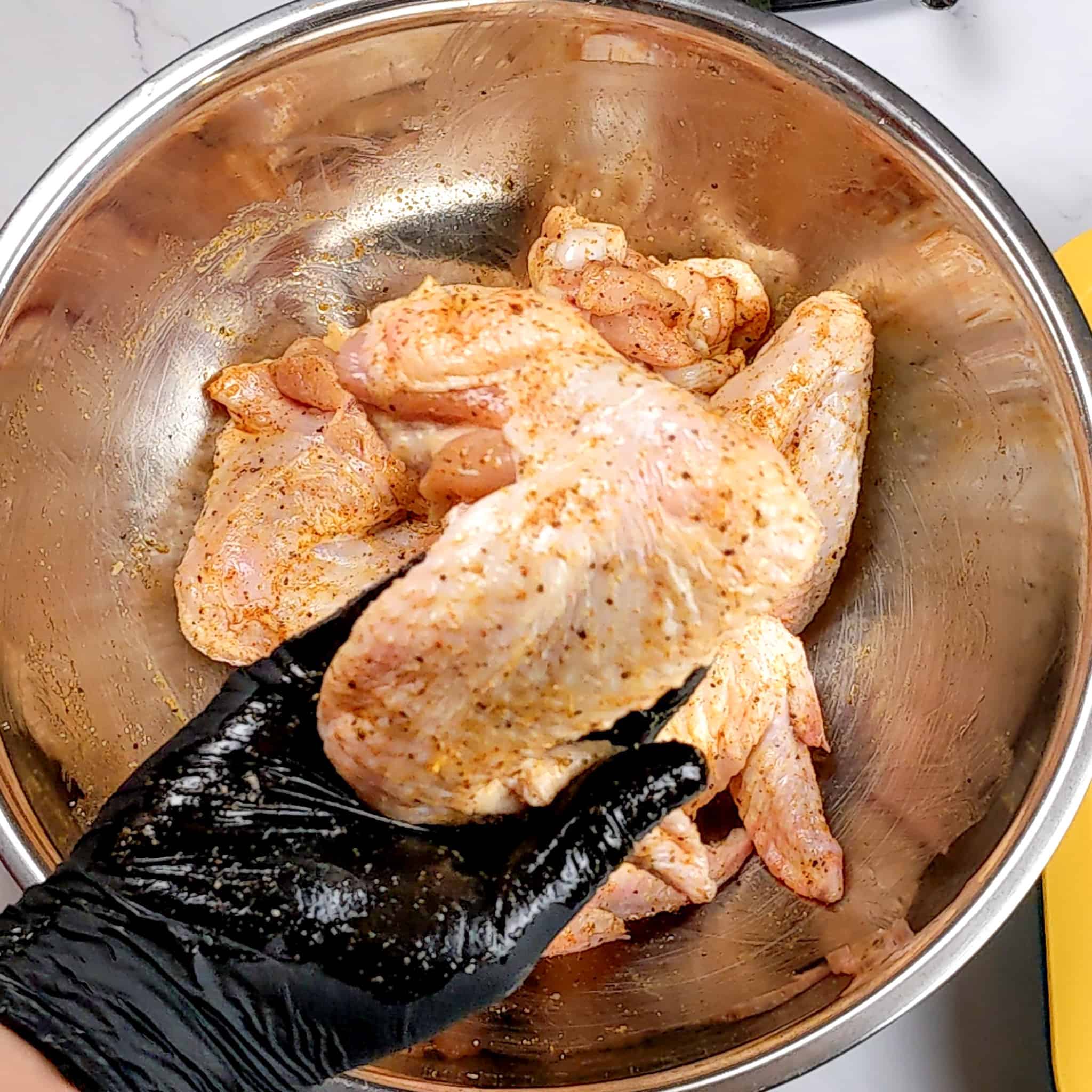 A raw seasoned whole chicken wing held in a gloved hand over a stainless steel bowl with the rest of the wings