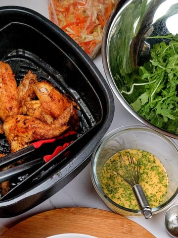 Starting at the top going clockwise, the shredded vegetables in the scotch bonnet pepper citrus vinaigrette in a resealable zipper bag, next to a stainless steel bowl with arugula, next to the finished vinaigrette with chopped herbs in a circle glass container with a mini whisk, next to the pulled out roasting pan and air fryer basket of the Ninja Foodi 5-in-1 Indoor Electric Grill with Air Fryer insert with the cooked seasoned whole chicken wings.