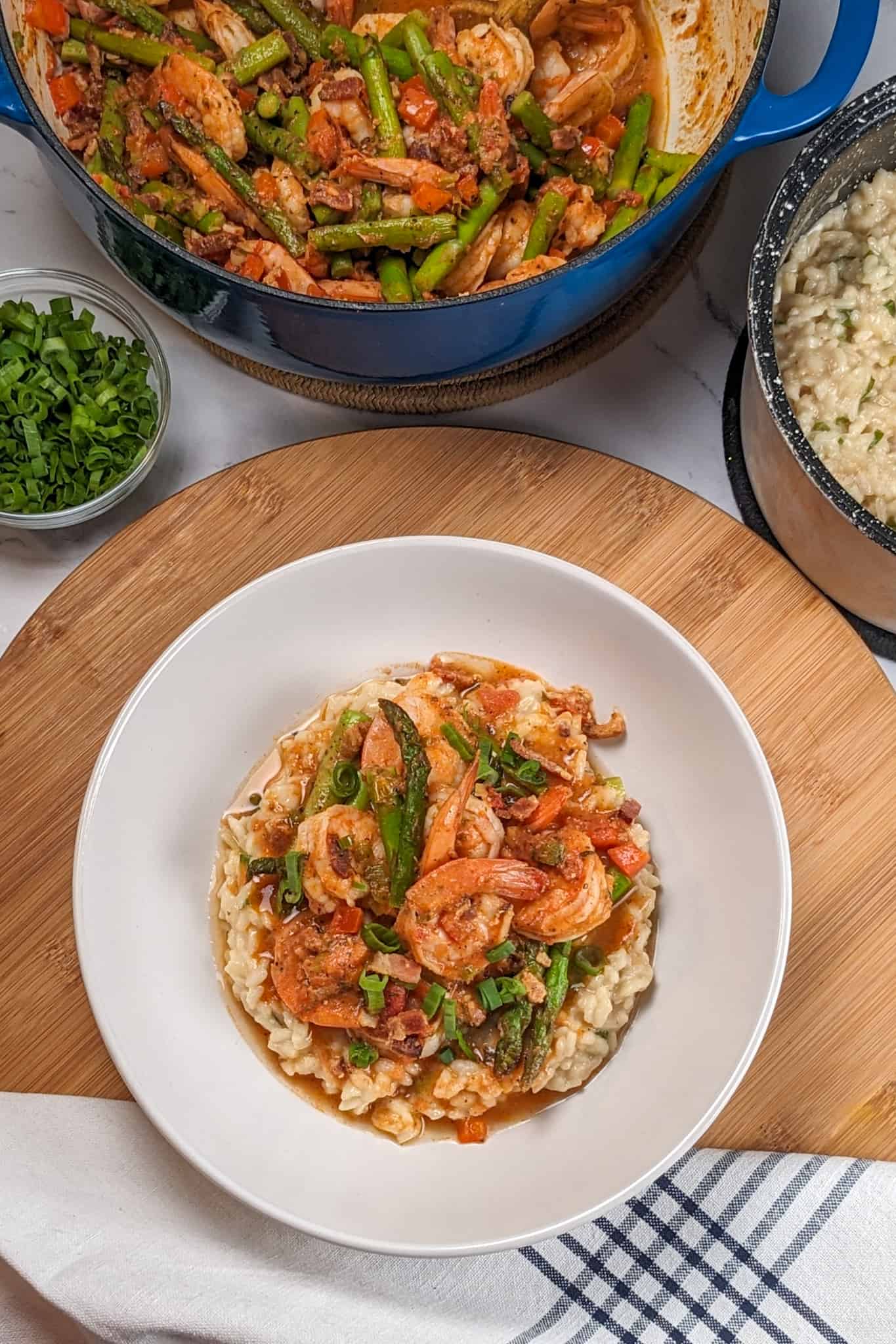 Top view of the Spicy Creole Shrimp and White Cheddar Cheese Risotto in a wide rim bowl on a wooden lazy susan next to a kitchen towel, surrounded by a small glass bowl of sliced scallions, a dutch oven of shrimp and asparagus and a canister of cooked risotto.