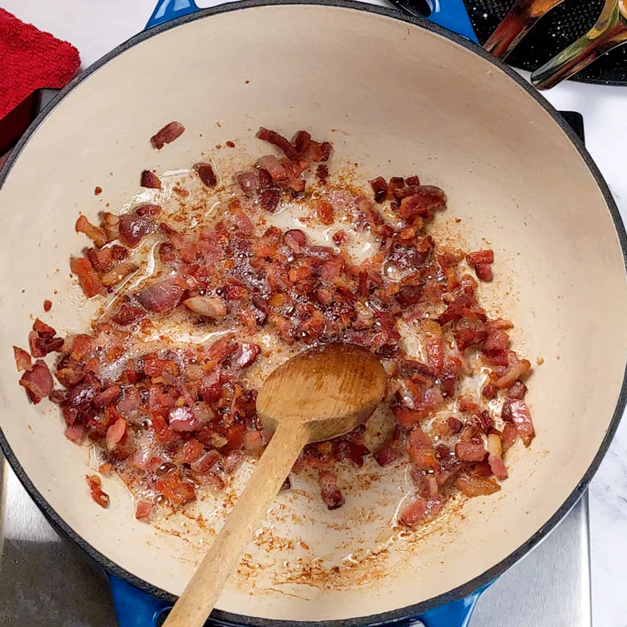 crispy cooked diced bacon being stirred in a enameled dutch oven with a wooden spoon