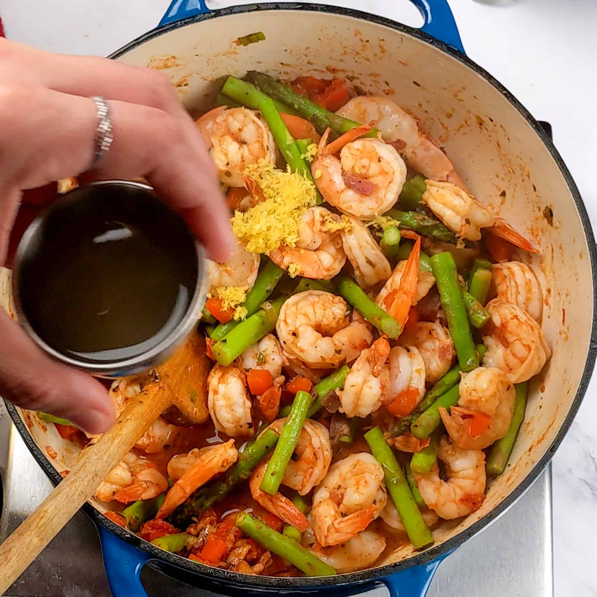 cooked diced red bell pepper with bacon, cooked sliced scallions, chopped garlic and tomato sauce, shrimp and asparagus cooked in a pot with a wooden spoon with a small cup of lemon juice held over the pot