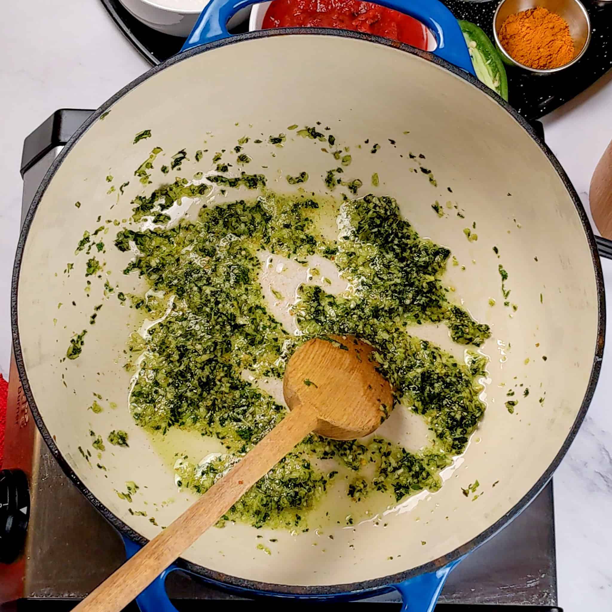 The blended aromatics of garlic, ginger and cilantro frying in oil and ghee in an lodge enameled Dutch oven for the Air Fryer Roasted Chicken Chickpeas Vegetable Curry recipe cooked with a wooden spoon resting in it.