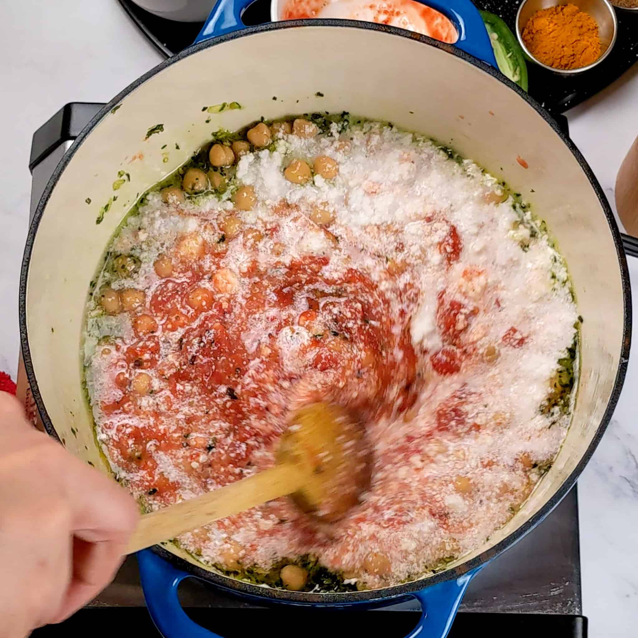 a wooden spoon stirring fire-roasted tomatoes, coconut milk, and chickpeas in the aromatics.