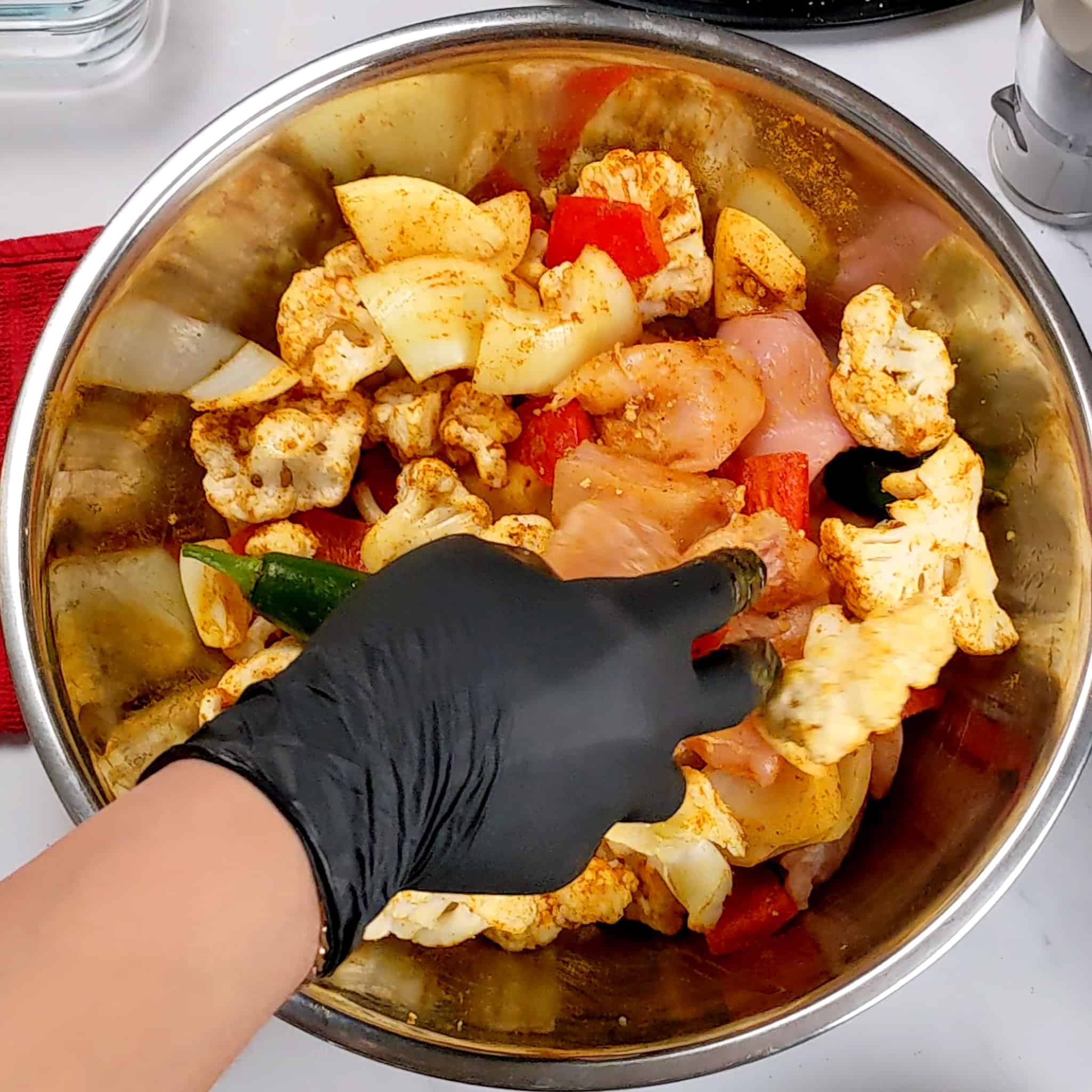 a gloved hand mixing the chicken breast, cauliflower, onions and red bell pepper in a large stainless steel mixing bowl for the Air Fryer Roasted Chicken Chickpeas Vegetable Curry recipe.