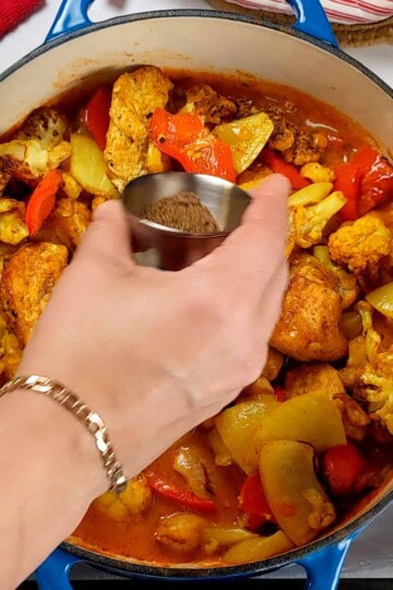 garam masala in a stainless steel condiment cup being poured into the chicken vegetable curry in the dutch oven.