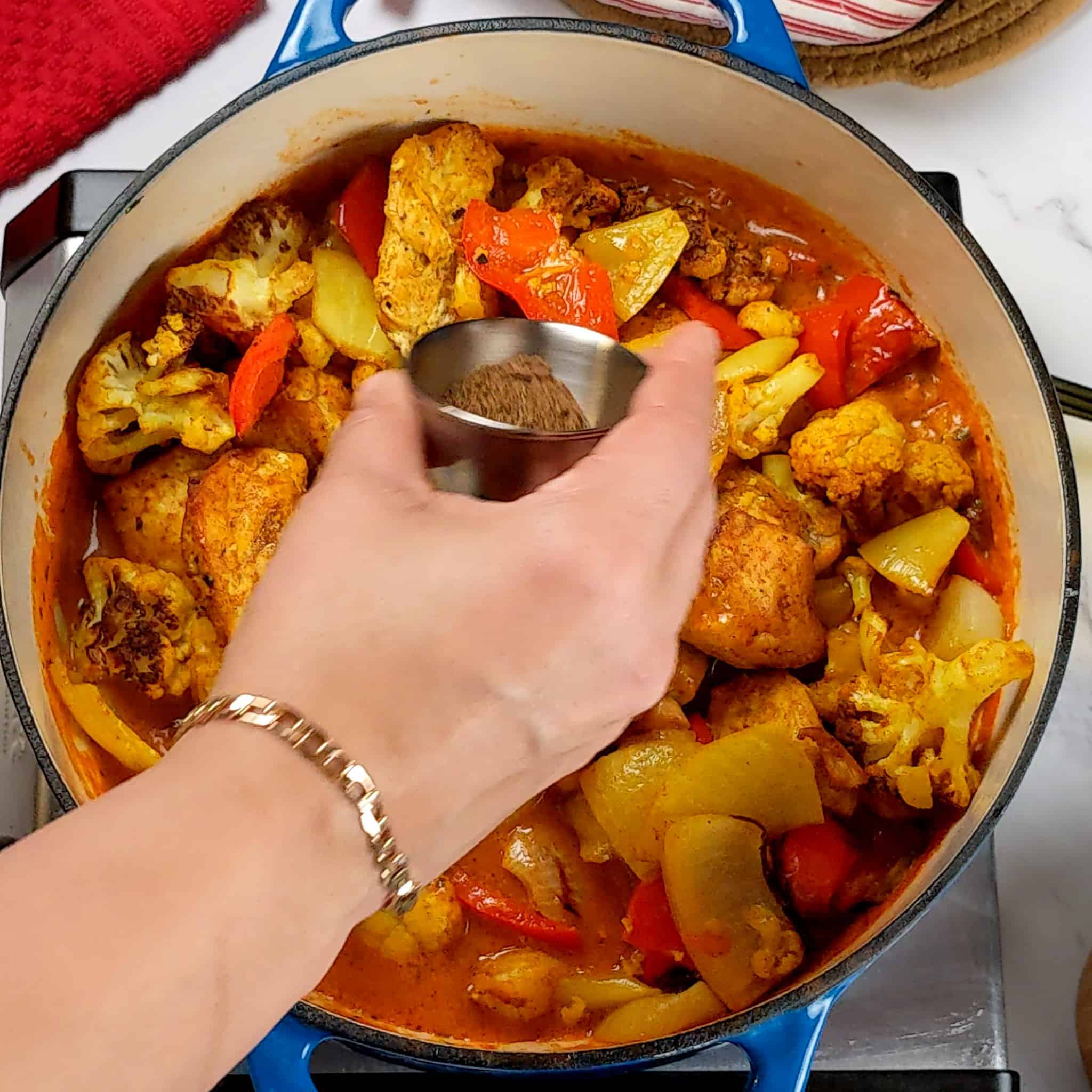 garam masala in a stainless steel condiment cup being poured into the chicken vegetable curry in the dutch oven.