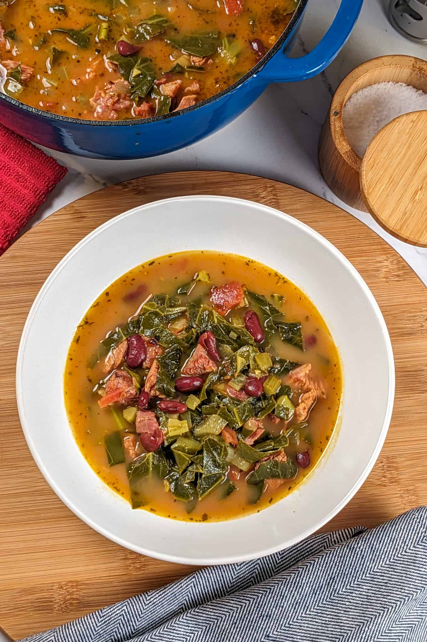 Top view of the Scotch Bonnet Smoked Turkey Collard Greens Beans Soup in a wide rim bowl surrounded by a kitchen towels a dutch oven with the remaining soup, and a salt cellar