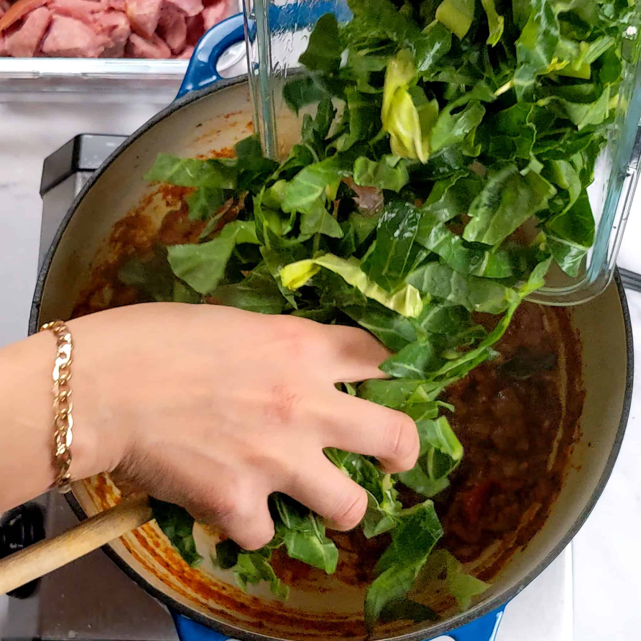 shredded collard greens in a glass rectangle container being poured into the dutch oven with the soup base with the help of a hand