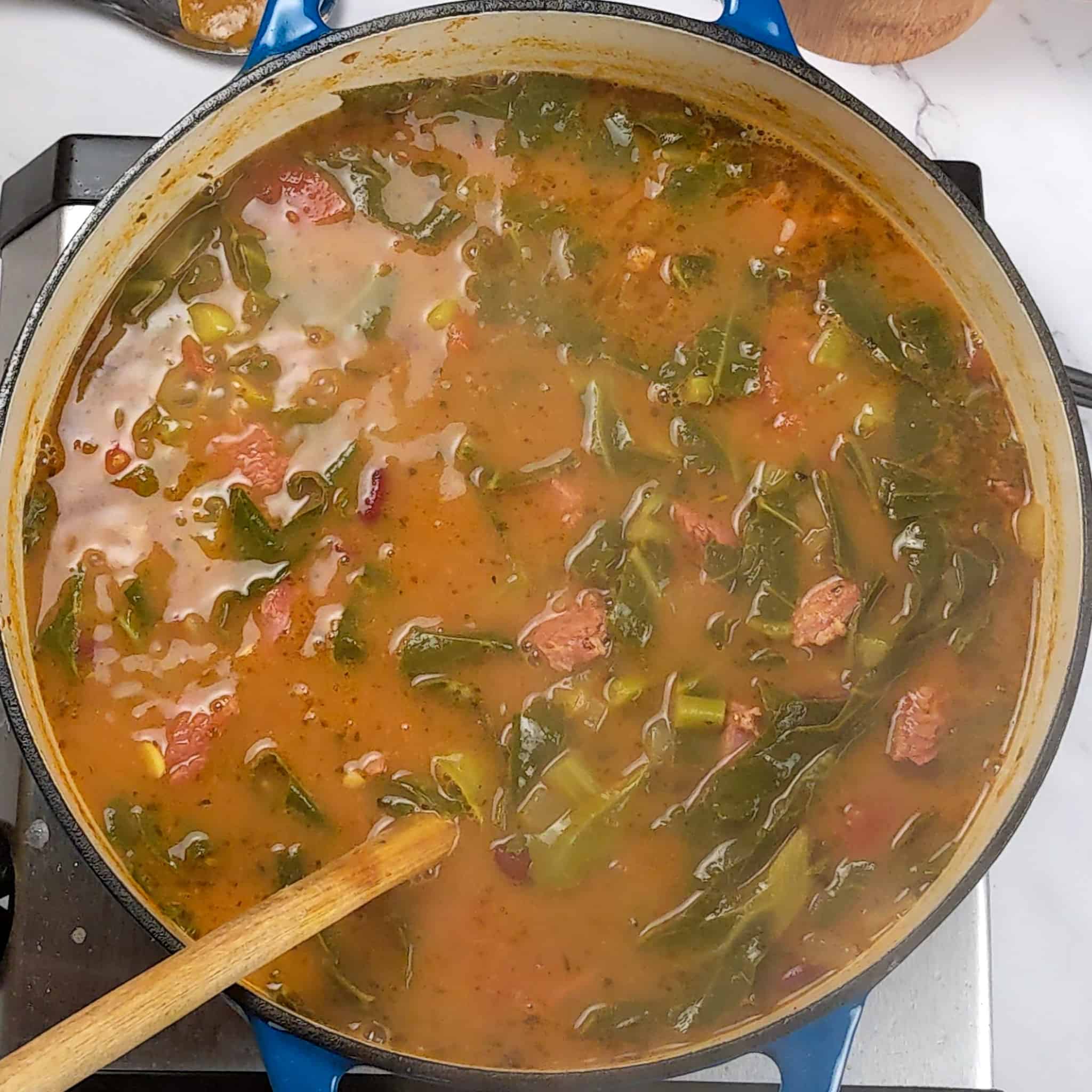 top view of the Scotch Bonnet Smoked Turkey Collard Greens Beans Soup in a lodge enameled dutch oven with a wooden spoon resting in it