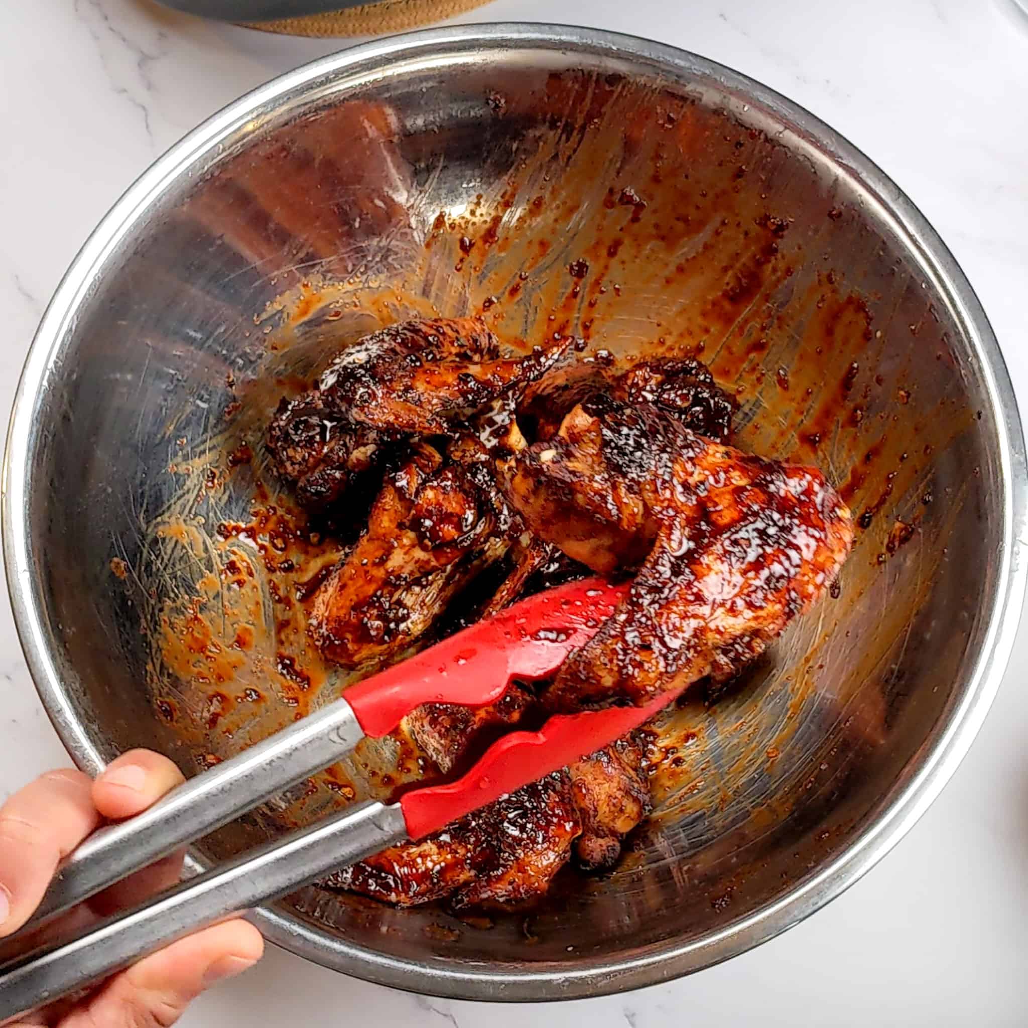 a pair of kitchenaid silicone tongs holding up a piece of the sticky date teriyaki glazed chicken wing over the rest of the wings that rest in a large stainless steel mixing bowl