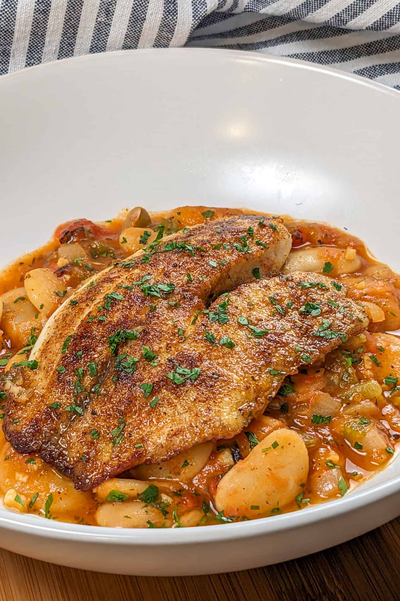 Top side view of Pan-Seared Cajun Spiced Tilapia on White Bean Stew recipe in a wide rim bowl next to a kitchen towel.