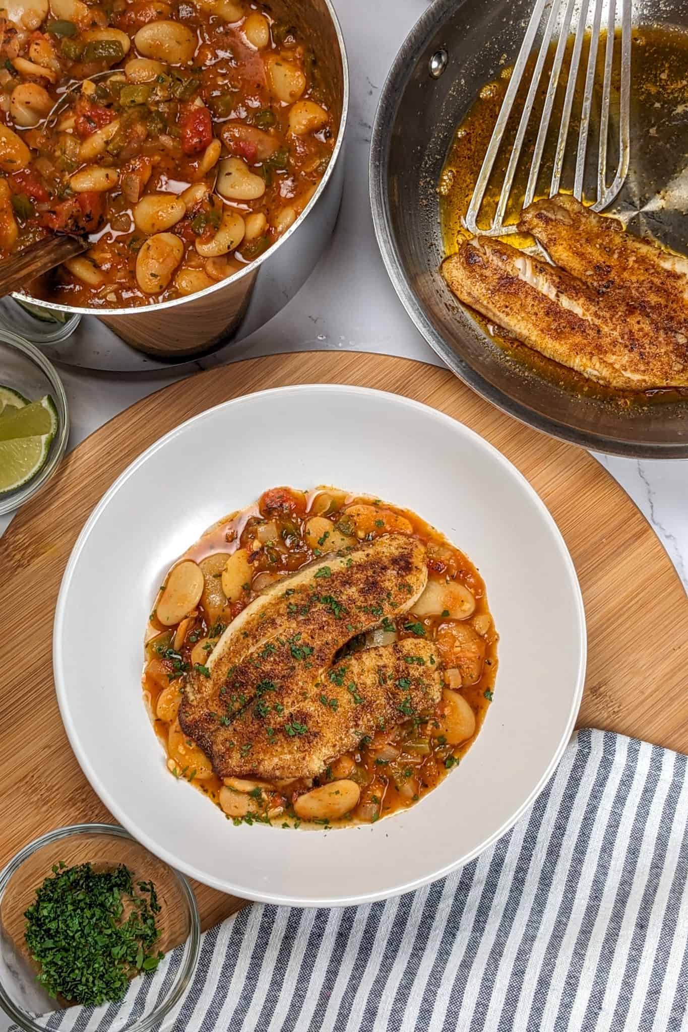 Top view of Pan-Seared Cajun Spiced Tilapia on White Bean Stew recipe in a wide rim bowl next to a kitchen towel surrounded by a small glass ramekin with fresh chopped parsley, another with lime wedges, and all-clad saucepan with white bean stew simmered in a tomato base sauce , and a all-clad stainless steel frying pan with a cajun spice crusted tilapia with a fish spatula resting next to it.