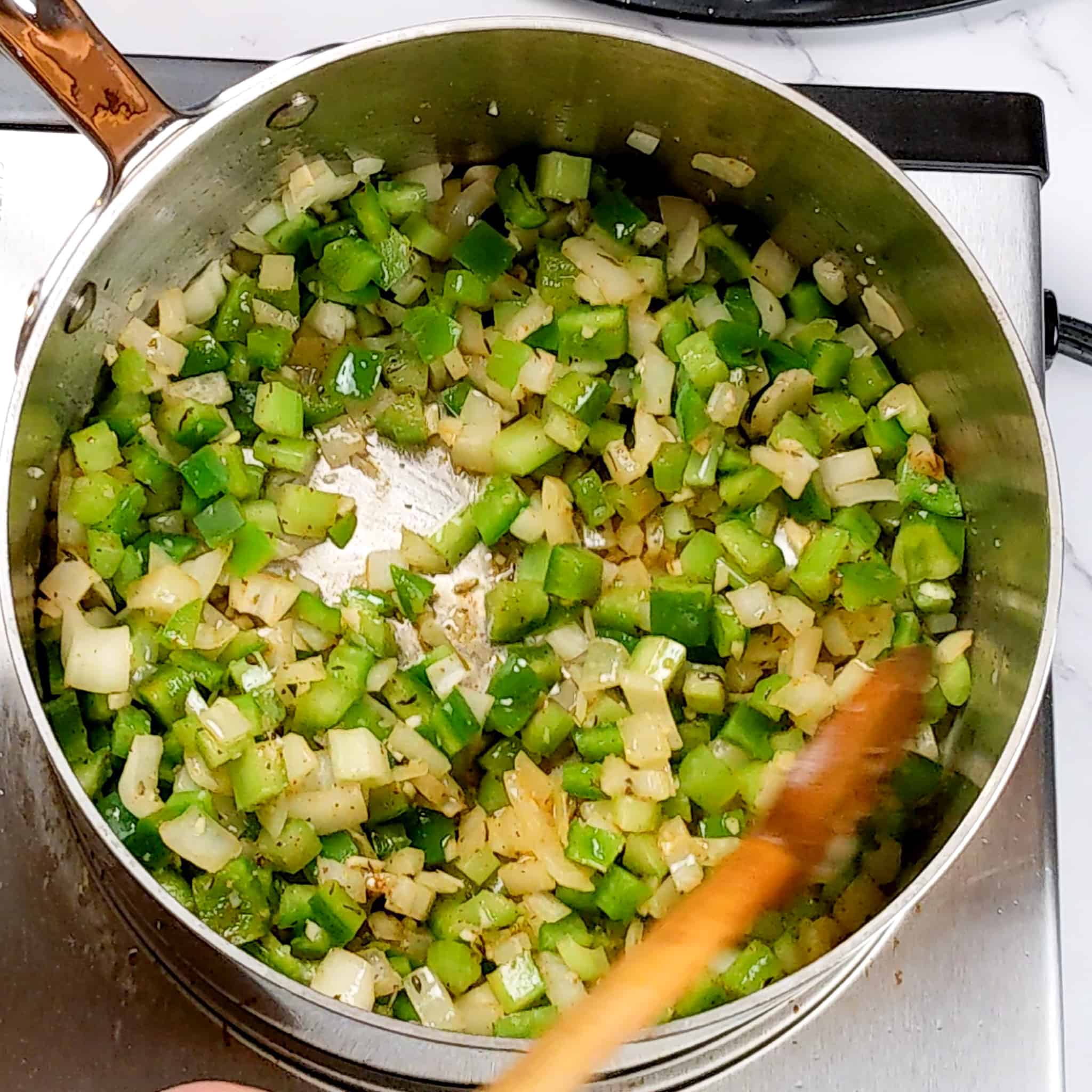 an all-clad stainless steel 3 quart sauce pan with diced onion, green bell pepper and celery sweating and being stirred with a wooden spoon.
