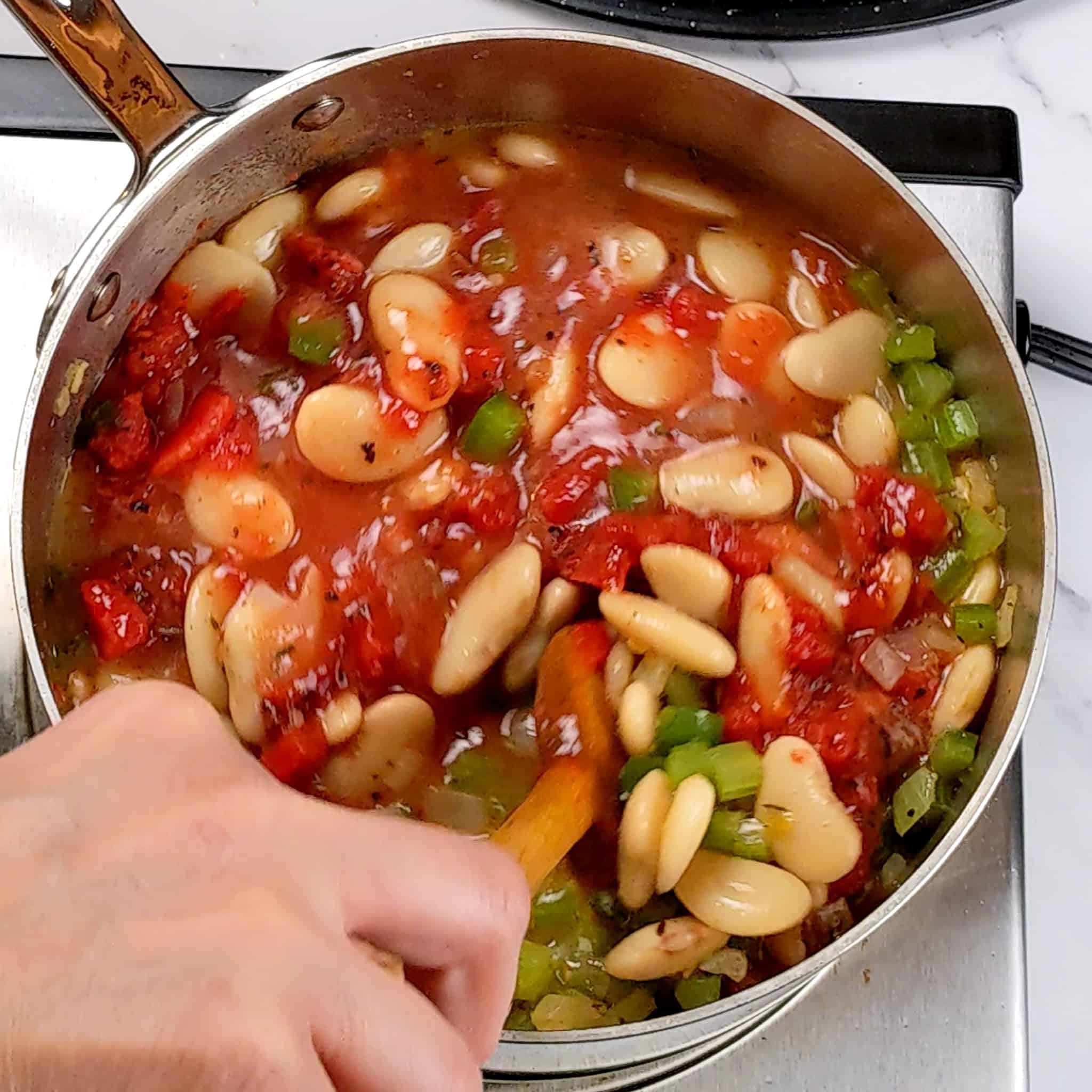 a saucepan of the holy trinity, tomato sauce and butter beans being stirred with a wooden spoon