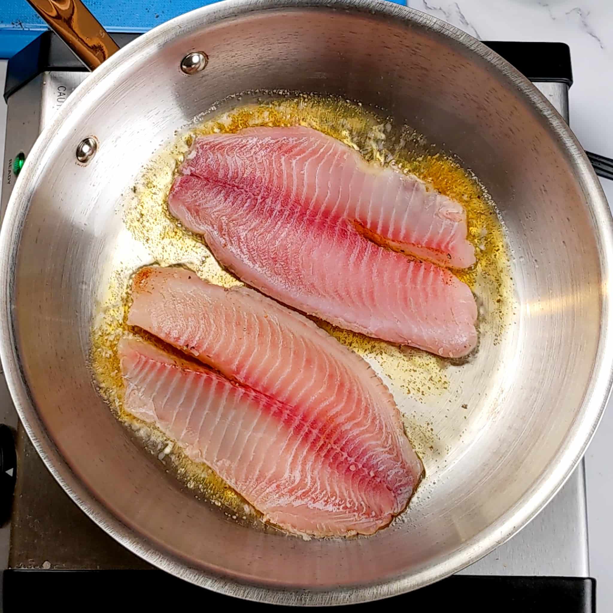 two tilapia filets season side down searing in an stainless steel frying pan.