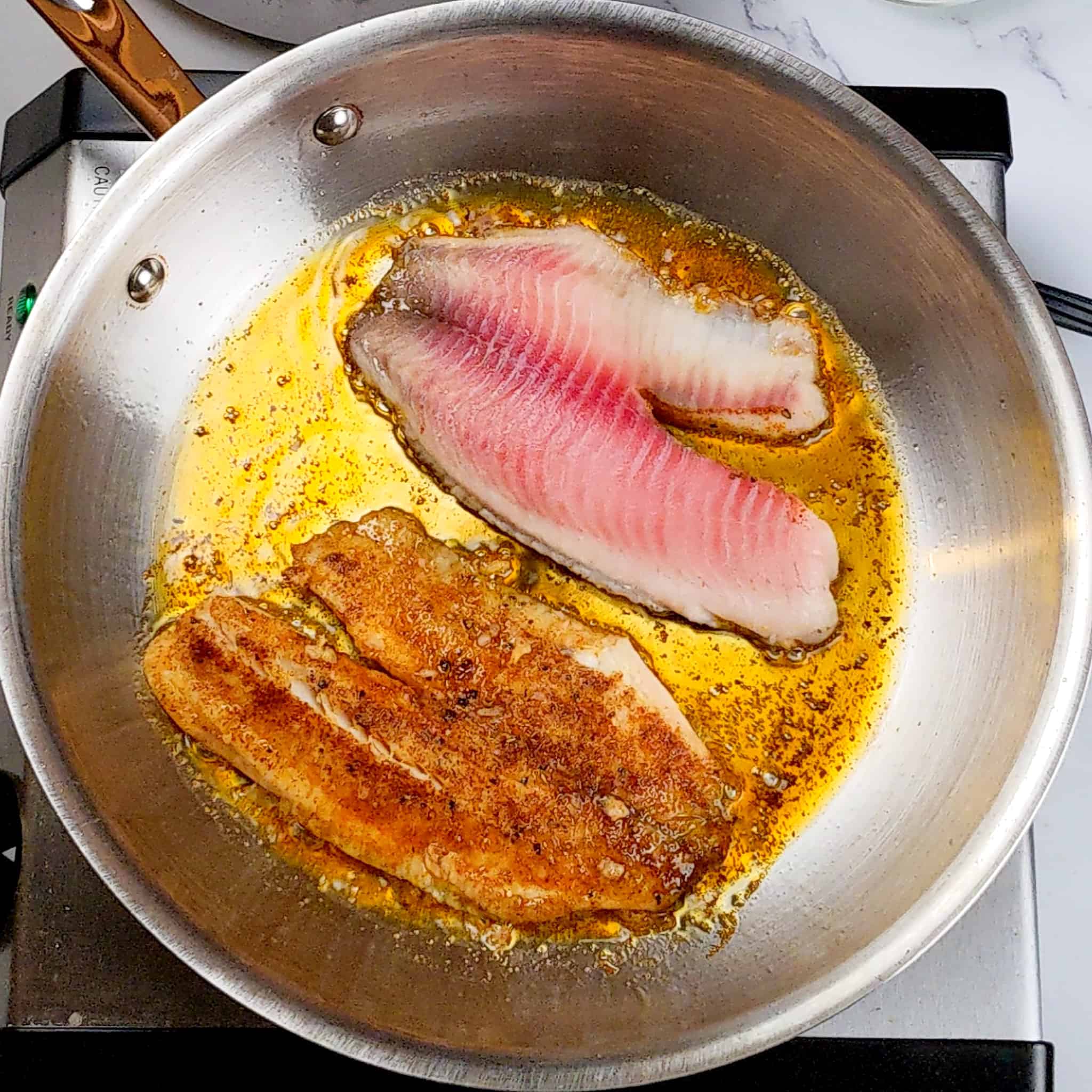 two tilapia filets, one season side down and the other flipped searing in an stainless steel frying pan.