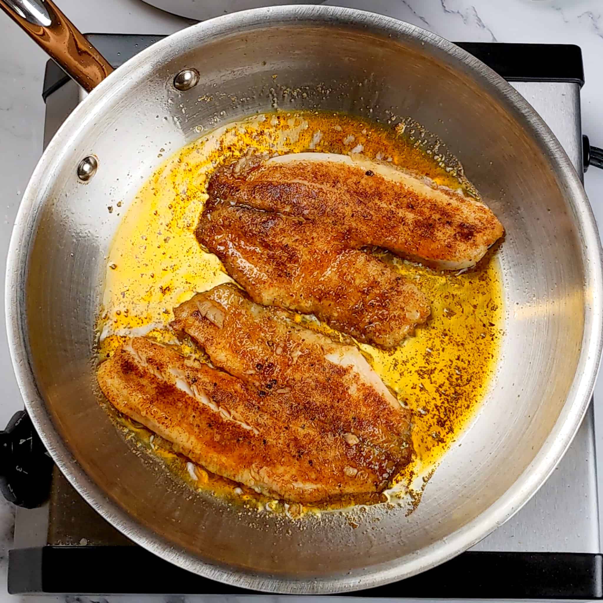 two tilapia filets, both flipped with season side up searing in an stainless steel frying pan.