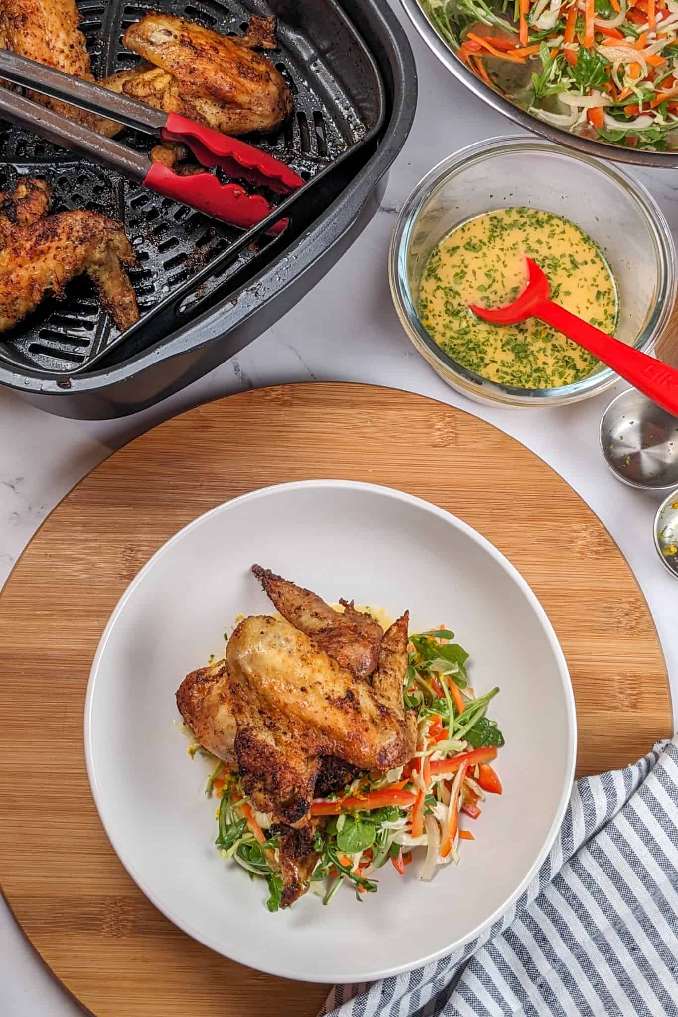 top view of the Best Air Fryer Caribbean Style Citrus Chicken Wings recipe in a wide rim bowl with salad on a lazy susan next to a kitchen towel, a container of the finished vinaigrette, salad and air fried chicken wings