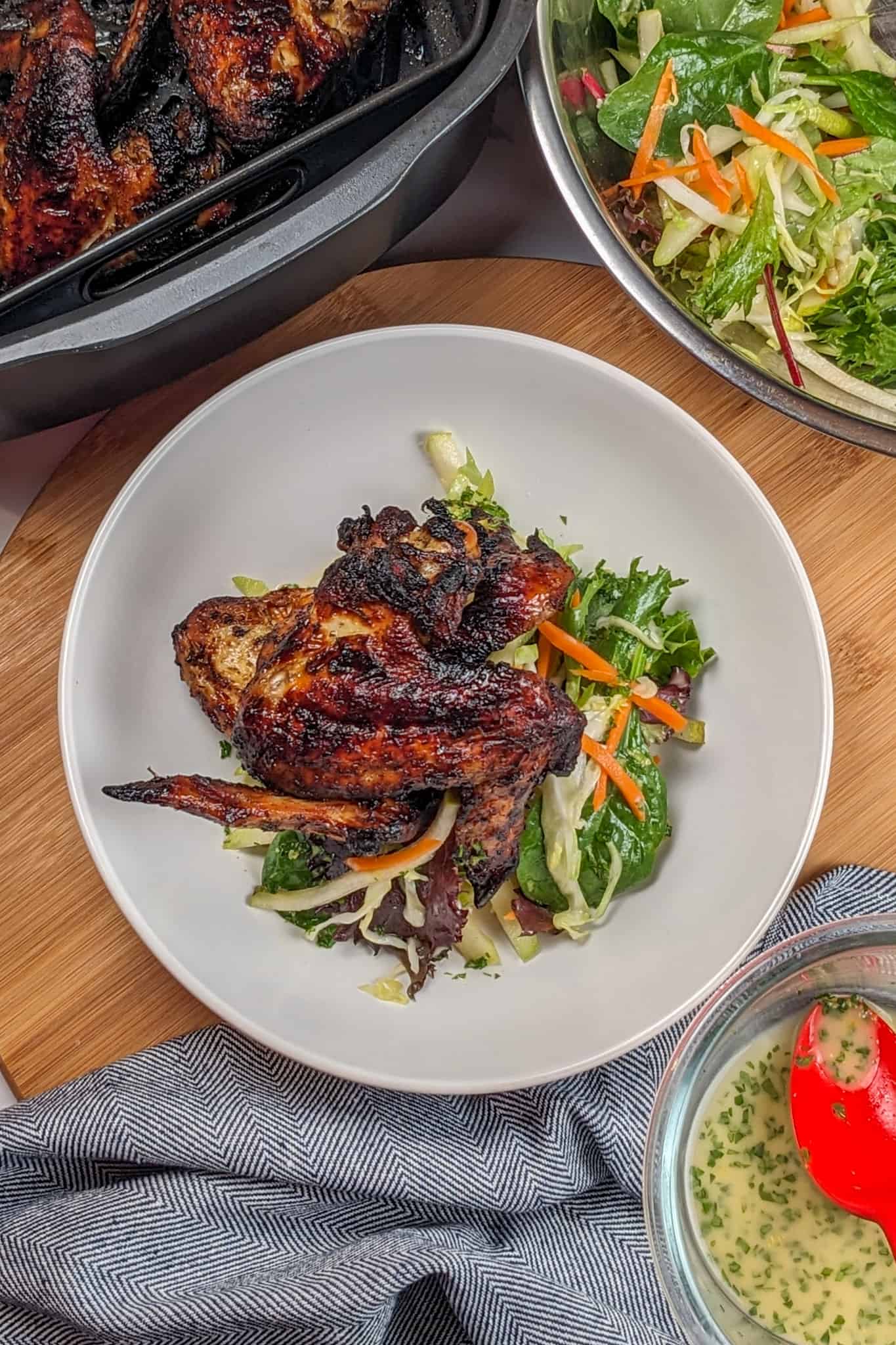 top view of the Crispy Air Fryer Honey Jerk Rubbed Chicken Wings recipe next to marinated vegetables with greens and vinaigrette in a wide rim bowl on a lazy susan next to a kitchen towel, glass bowl of vinaigrette with a silicone table spoon, next to a large stainless steel mixing bowl with the rest of the salad mixture , which sits next to an air fryer roasting pan with the air frying basket with the rest of the cooked honey jerk rubbed chicken wings.