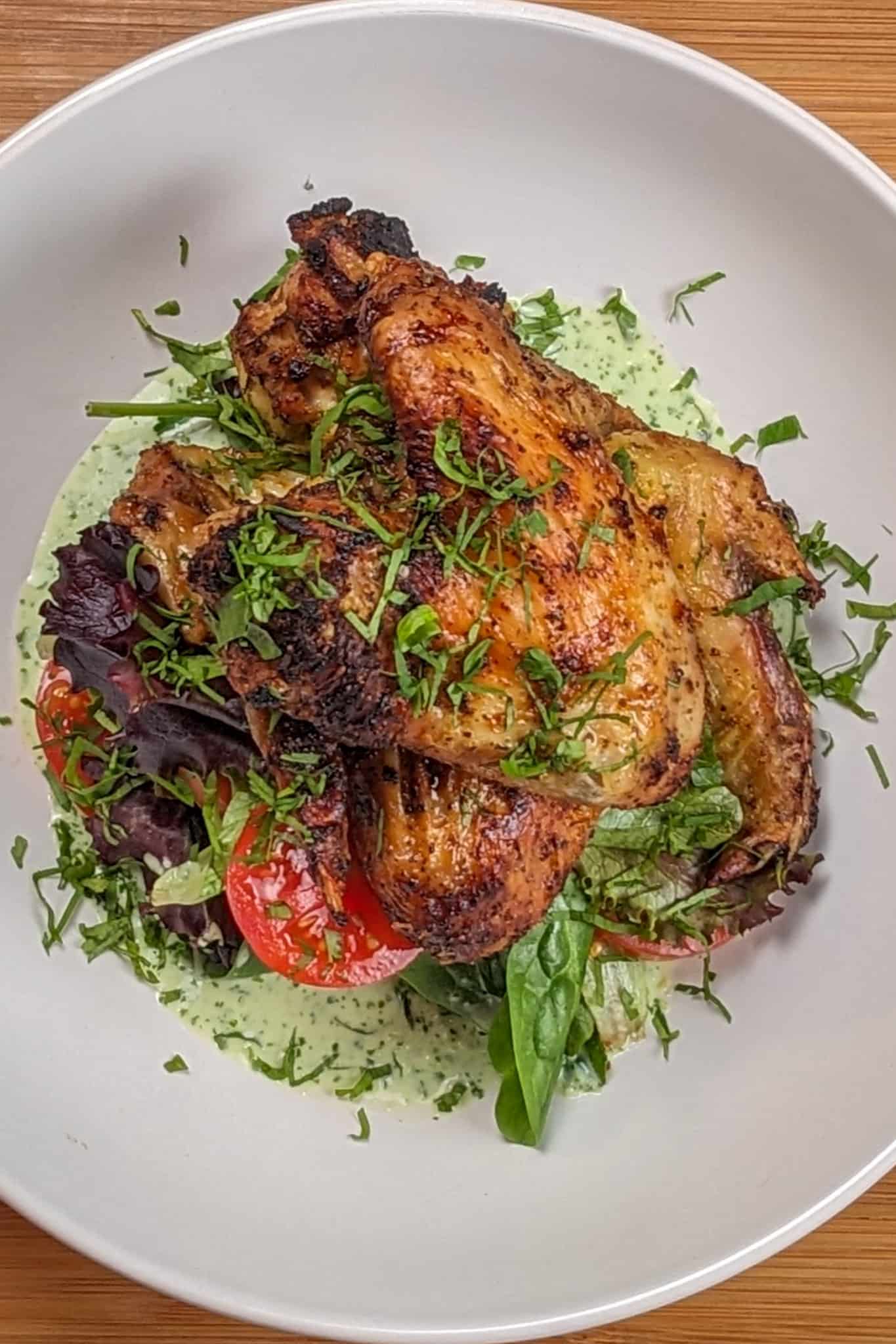 top view Plated Quick Air Fryer Lime Chicken Wings on greens with sliced campari tomatoes with Spicy Green Sauce on the bottom and the whole dish garnished with chiffonade herbs in a wide rim bowl