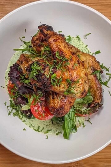 top view Plated Quick Air Fryer Lime Chicken Wings on greens with sliced campari tomatoes with Spicy Green Sauce on the bottom and the whole dish garnished with chiffonade herbs in a wide rim bowl