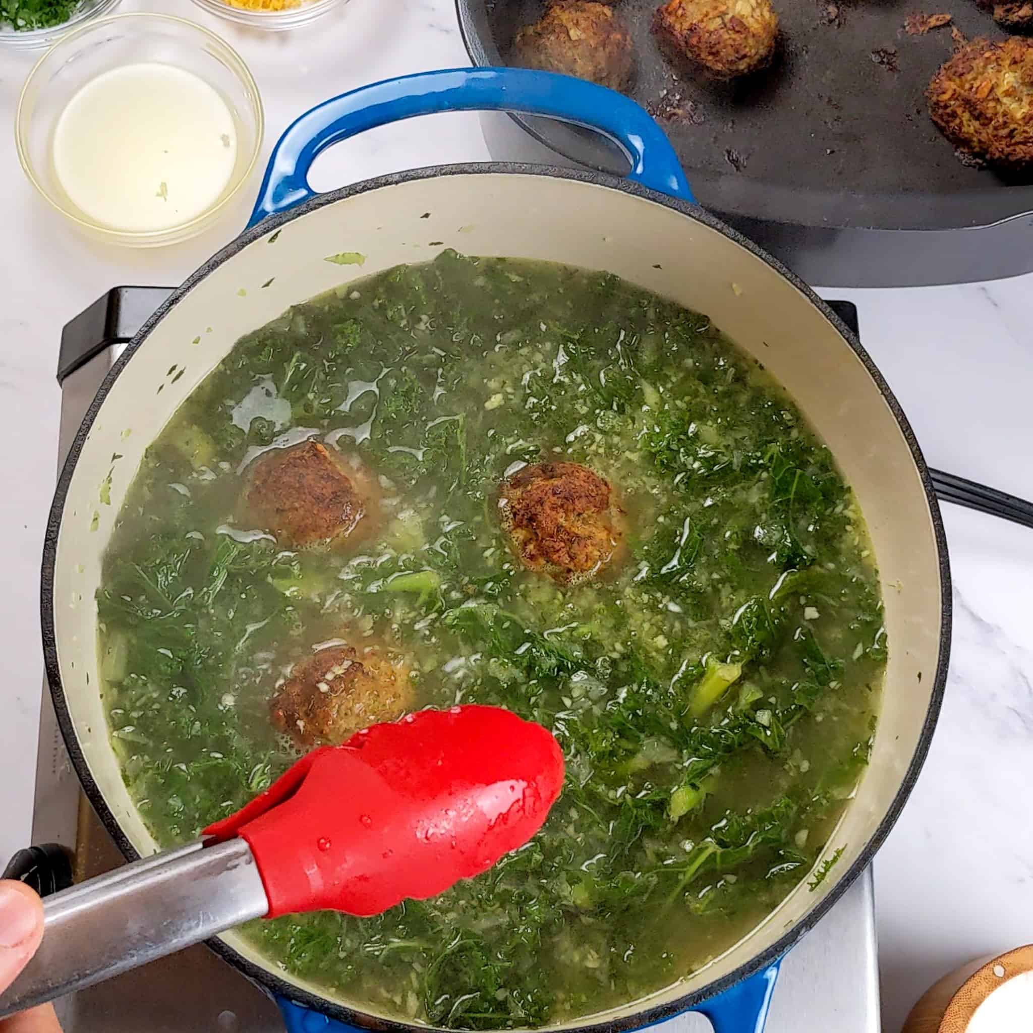 meatballs being added to the kale broth with a pair of kitchenaid silicone tongs for the Easy and Healthy Spicy Turkey Meatball Kale Soup