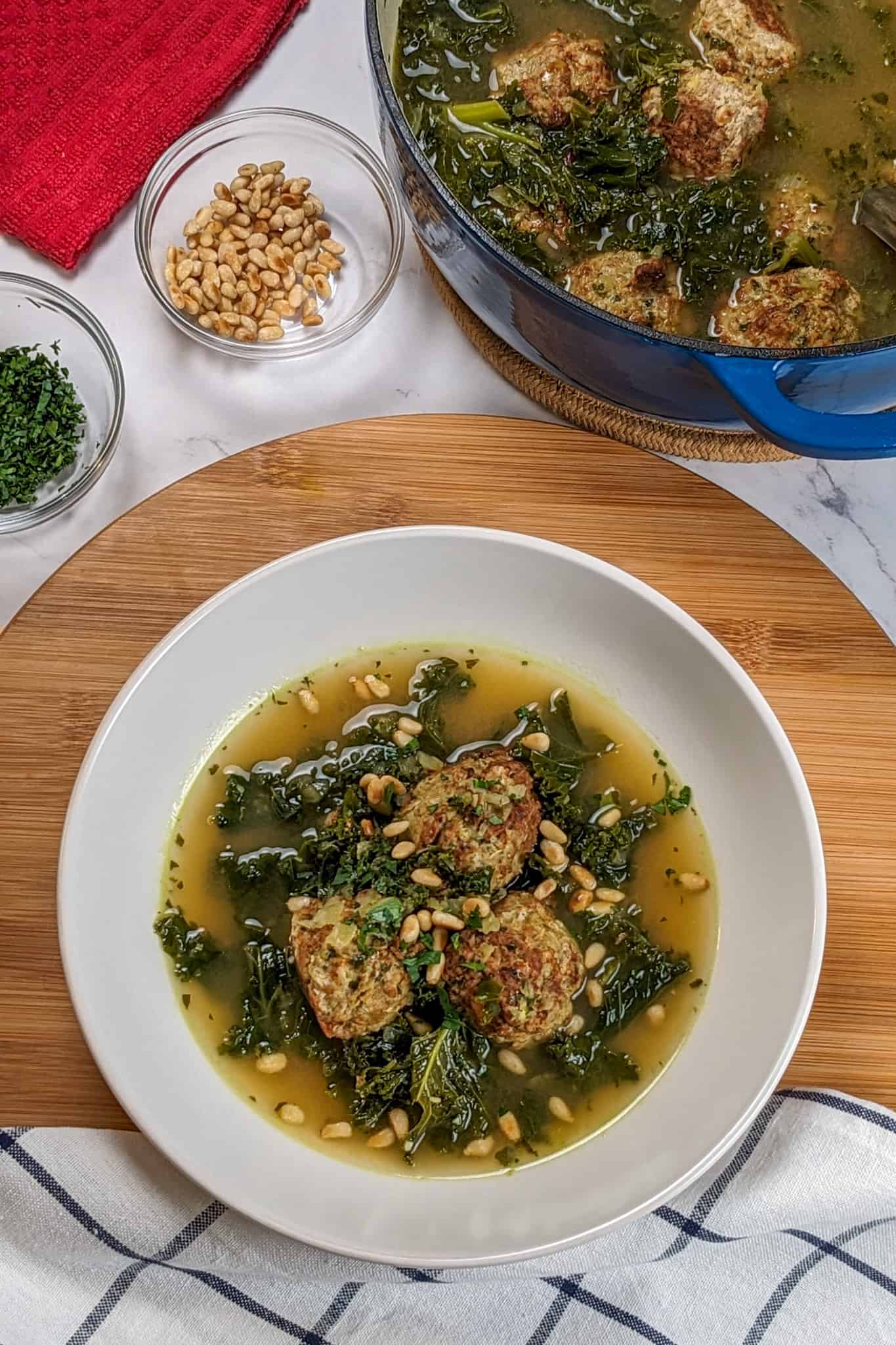 top close up view of the Easy and Healthy Spicy Turkey Meatball Kale Soup in a wide rim bowl garnished with toasted pine nuts surrounded by a container of pine nuts and chopped parsley separately in small glass bowls next to a dtuch oven of soup