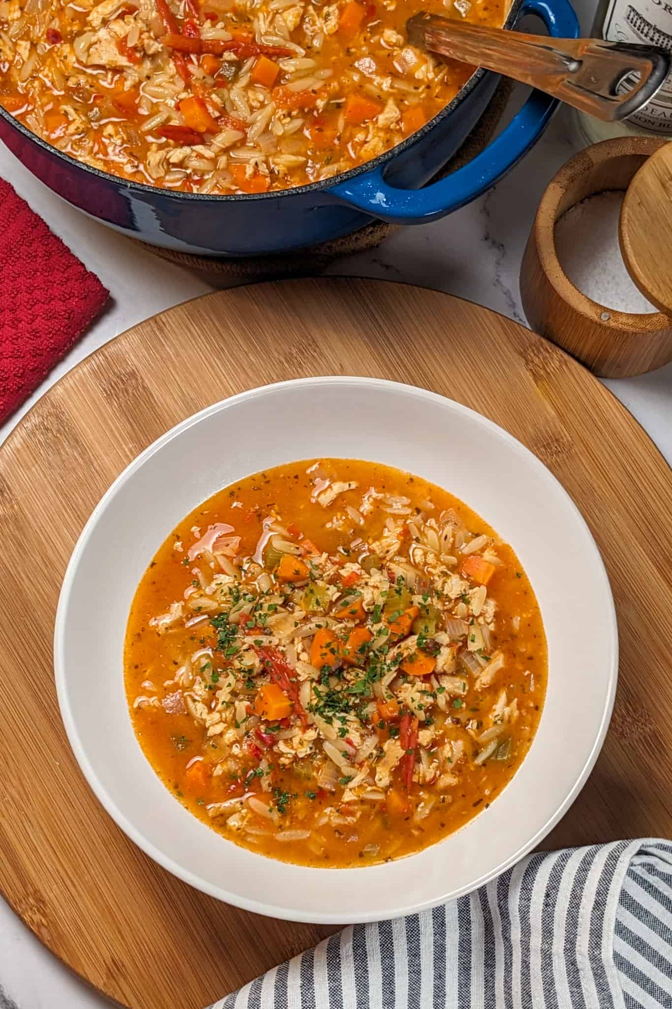 top view of the Lemon Calabrian Chili Chicken Orzo Soup in a wide rim bowl on a wooden lazy susan next to a kitchen towel, surrounded by an enameled Dutch oven with a ladle and salt cellar partially opened.