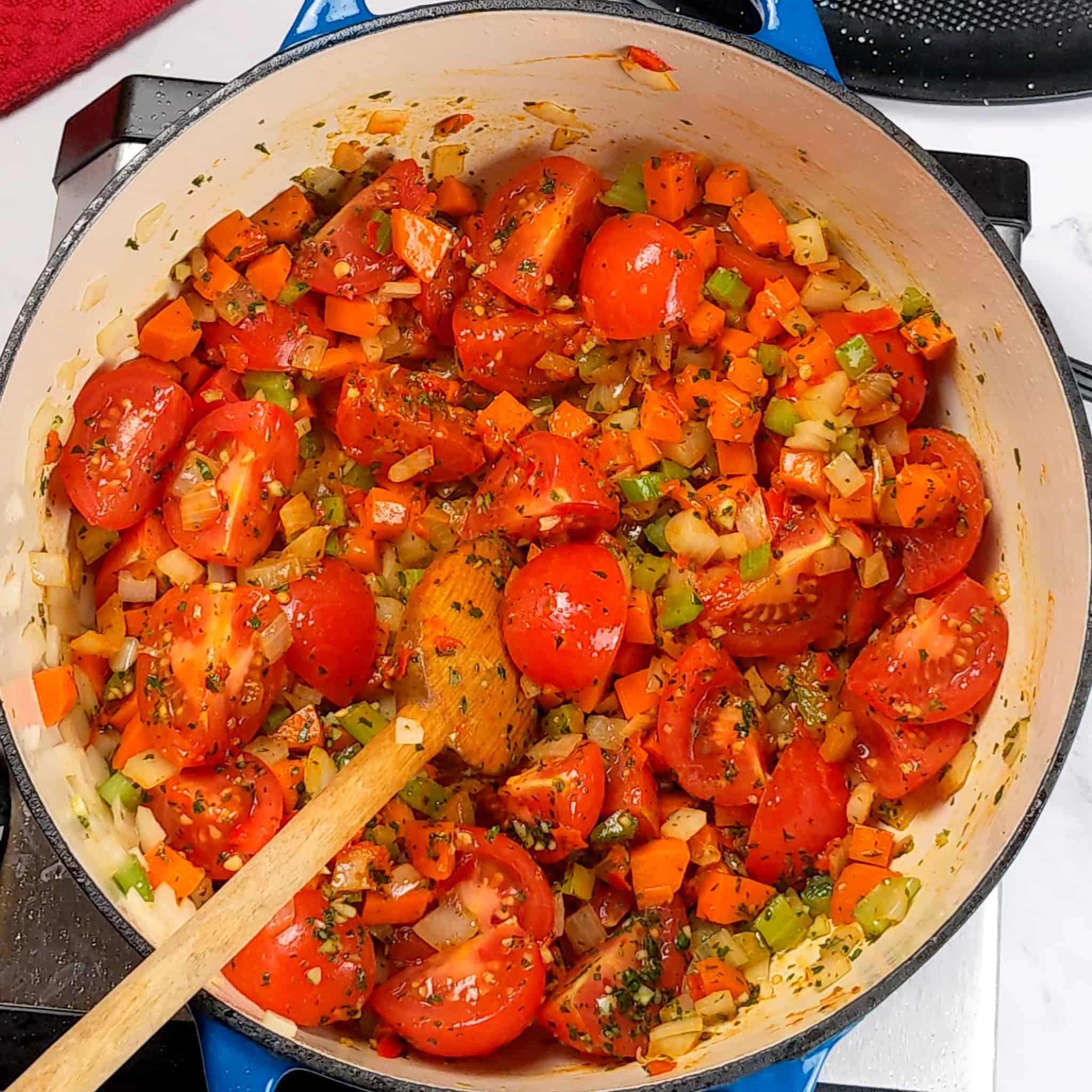 quartered campari tomatoes mixed in with herbs and mirepoix in dutch oven with a wooden spoon laying in it.
