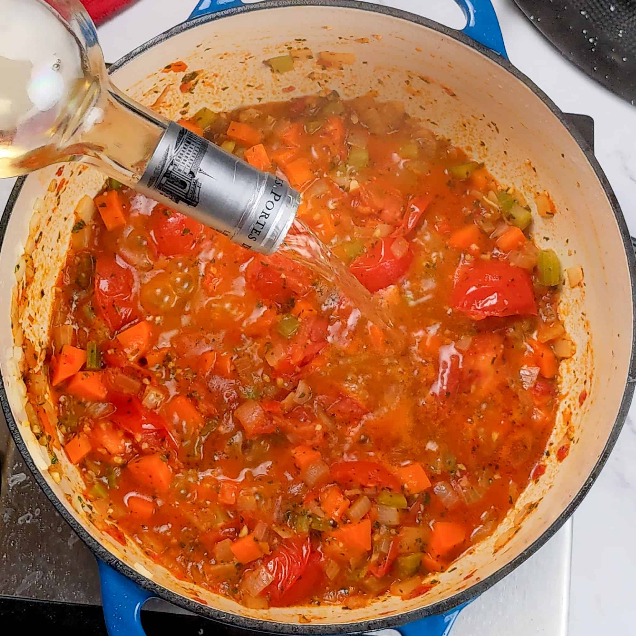 white wine being poured into cooked down campari tomatoes mixed in with herbs and mirepoix in an enameled dutch oven.