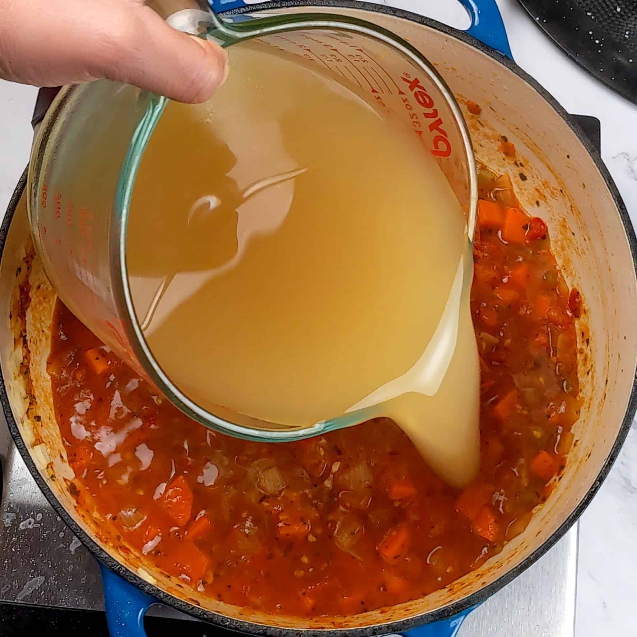 low-sodium chicken broth being poured into cooked down campari tomatoes mixed in with herbs and mirepoix in an enameled dutch oven.