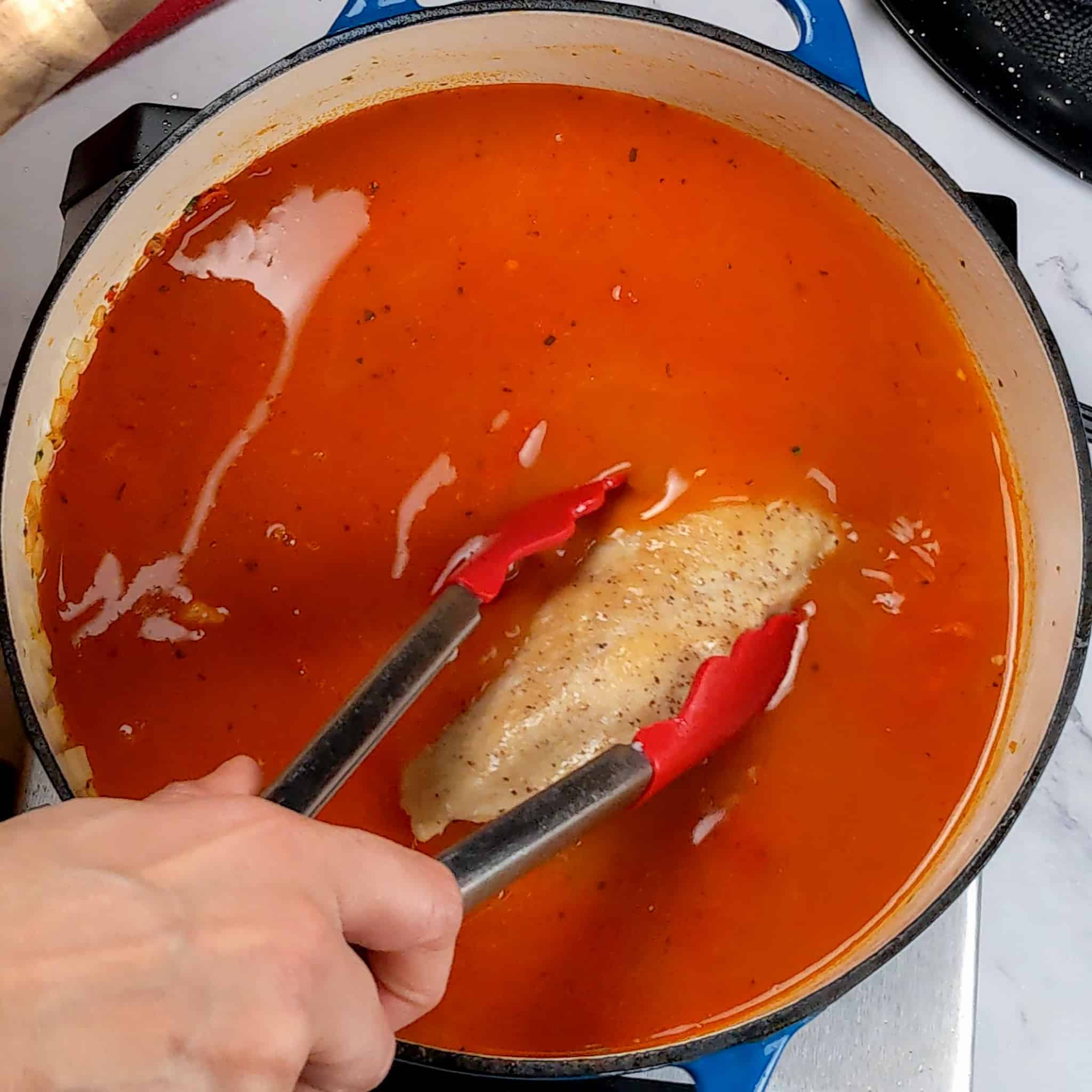 seared chicken breast being placed into a tomato based broth with a pair of kitchenaid silicone tipped tongs