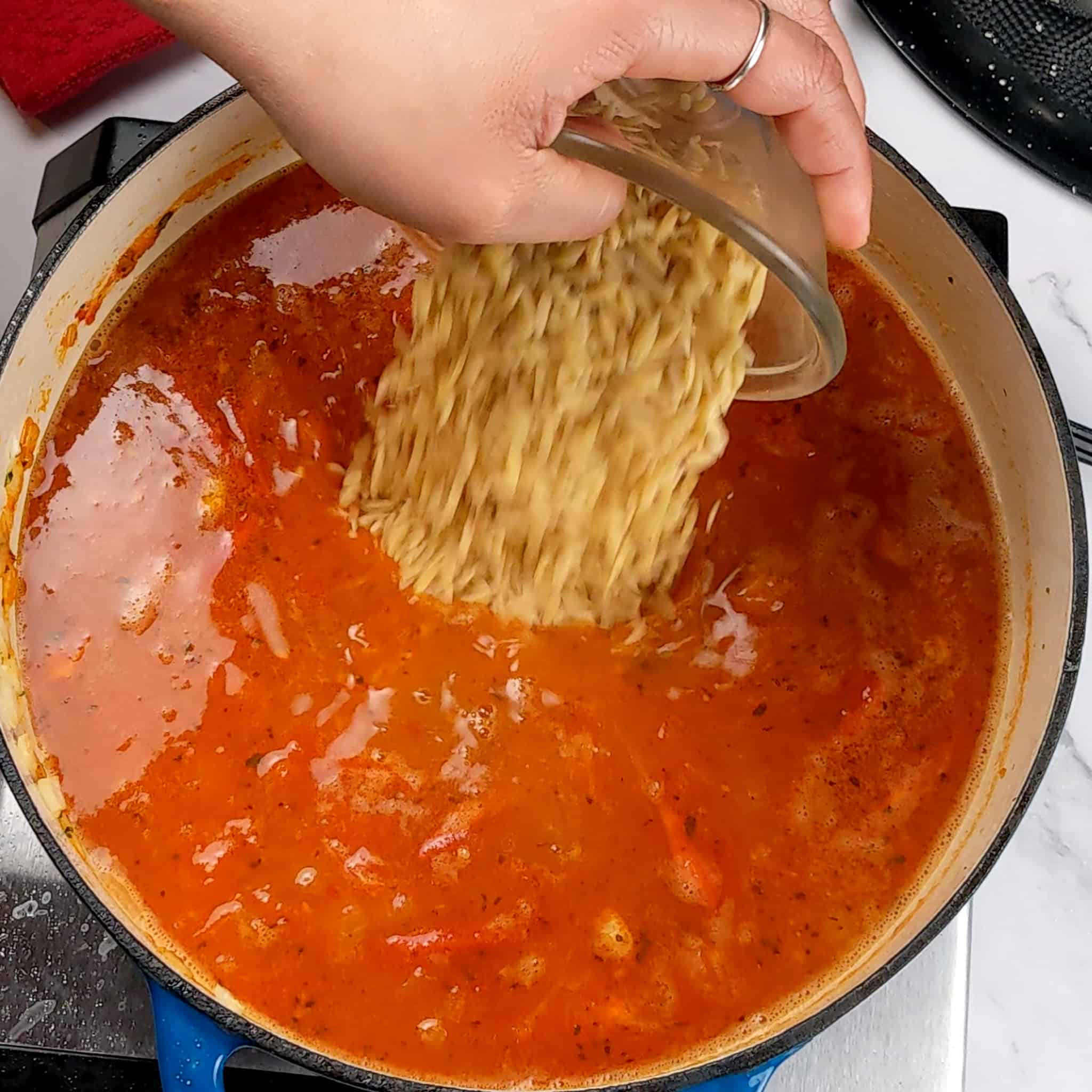dry orzo pasta being poured into a tomato base broth