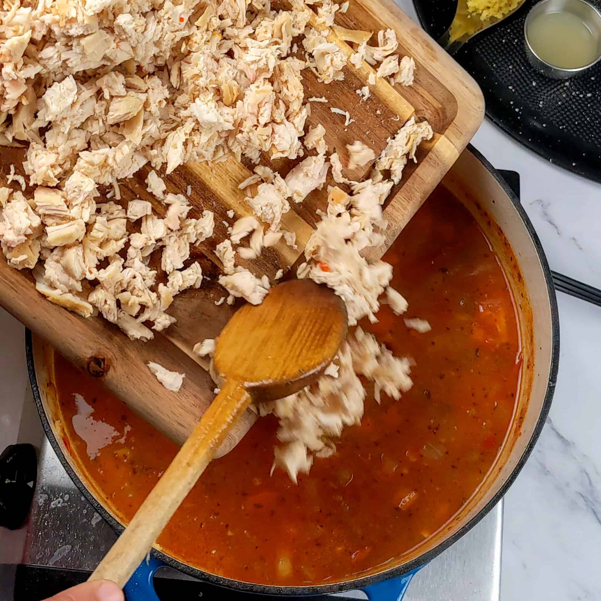 chopped boneless skinless cooked chicken breast on a small wooden cutting board being added to a tomato based broth with a wooden spoon