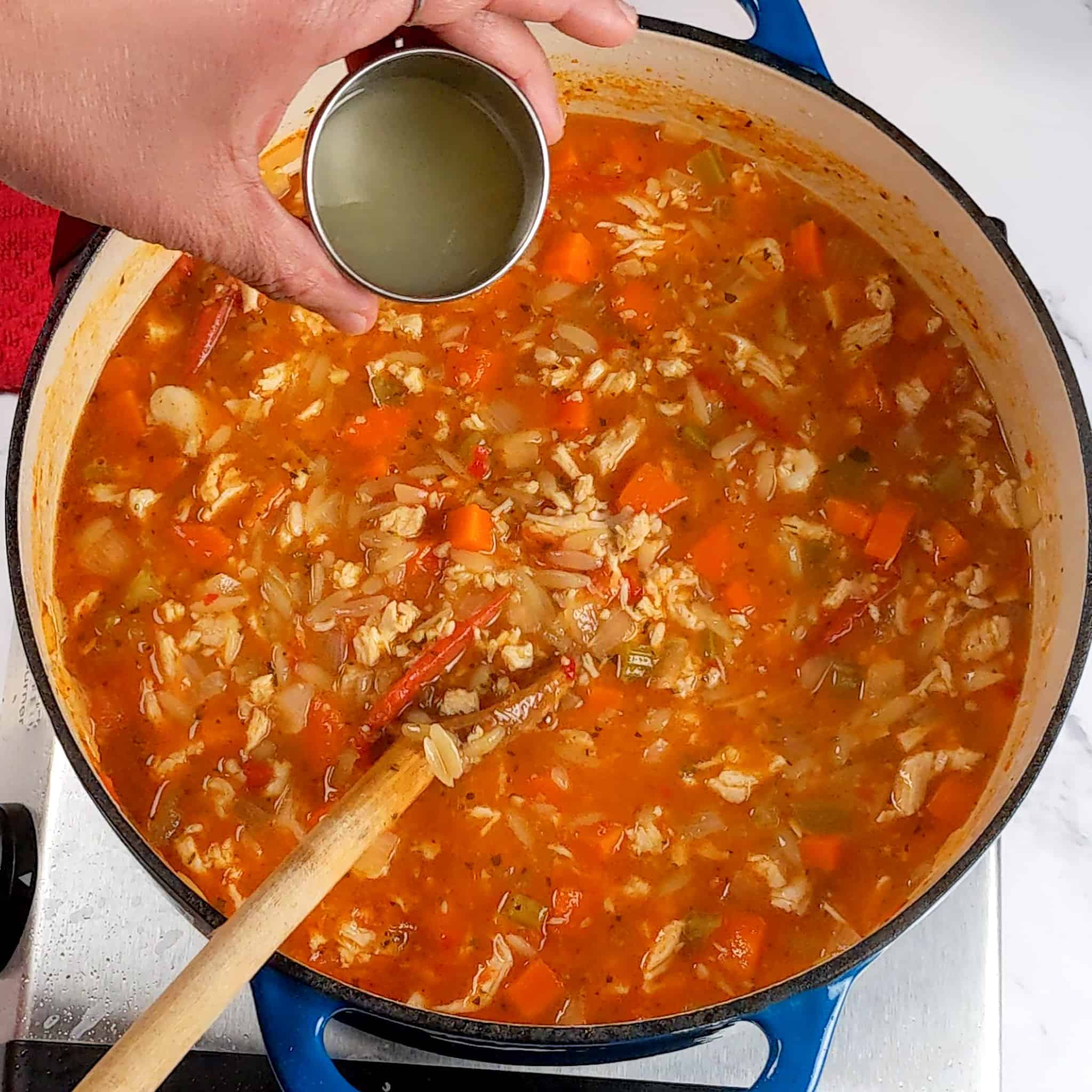 a hand holding a metal ramekin with lemon juice over the Lemon Calabrian Chili Chicken Orzo Soup in an enameled lodge dutch oven that has a ladle resting in it