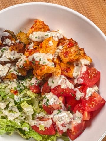 top view of the Air Fryer Calabrian Chili Salmon and Vegetables Bowl in a wide rim bowl, a dish where the cubed baked salmon, quartered campari tomatoes, shredded romaine lettuce and roasted vegetables share equal pie part with the rosemary tahini dressing drizzled in a zig zag on top