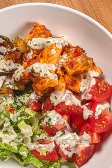 top view of the Air Fryer Calabrian Chili Salmon and Vegetables Bowl in a wide rim bowl, a dish where the cubed baked salmon, quartered campari tomatoes, shredded romaine lettuce and roasted vegetables share equal pie part with the rosemary tahini dressing drizzled in a zig zag on top