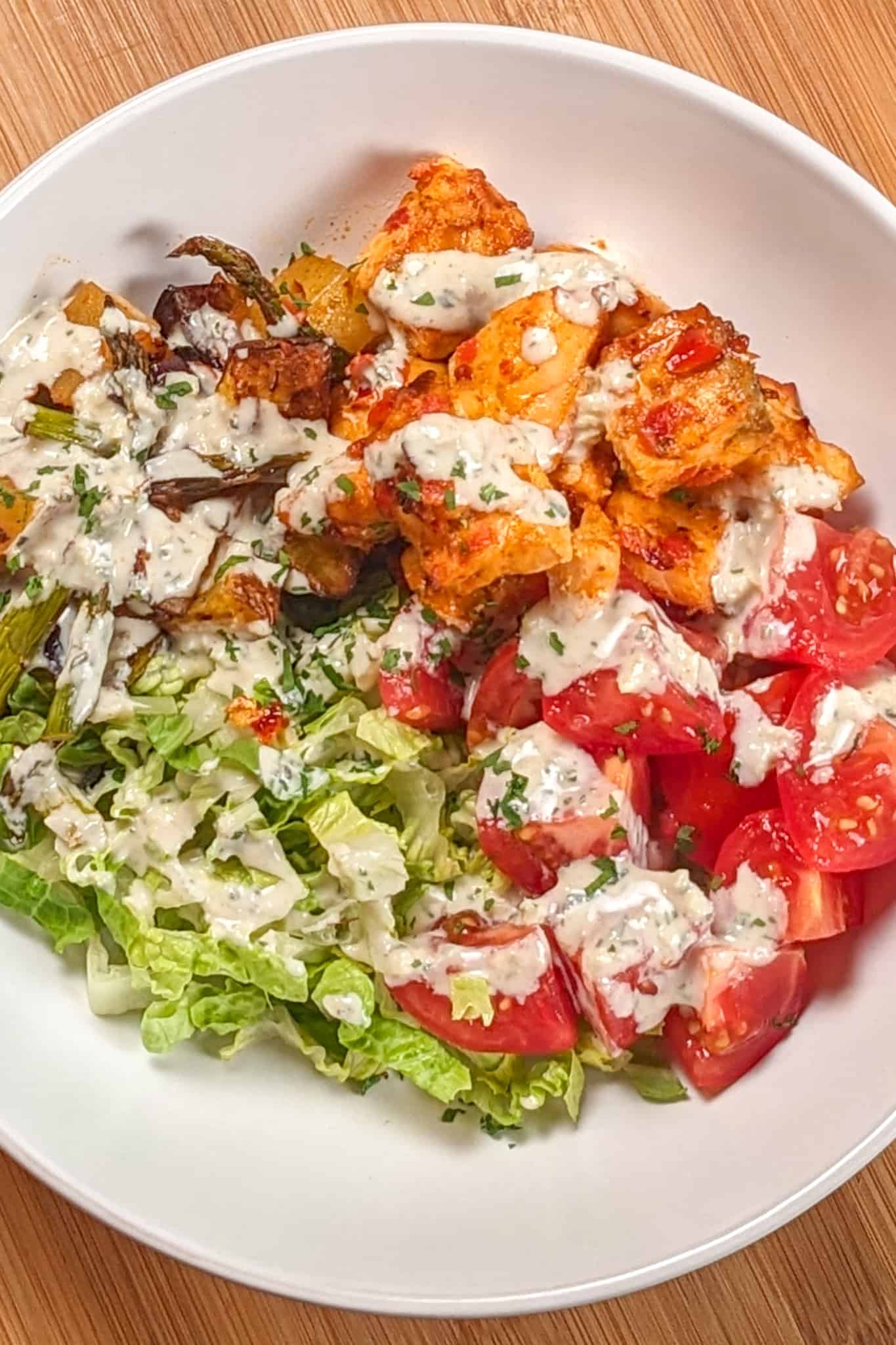 top view of the Air Fryer Calabrian Chili Salmon and Vegetables Bowl in a wide rim bowl, a dish where the cubed baked salmon, quartered campari tomatoes, shredded romaine lettuce and roasted vegetables share equal pie part with the rosemary tahini dressing drizzled in a zig zag on top