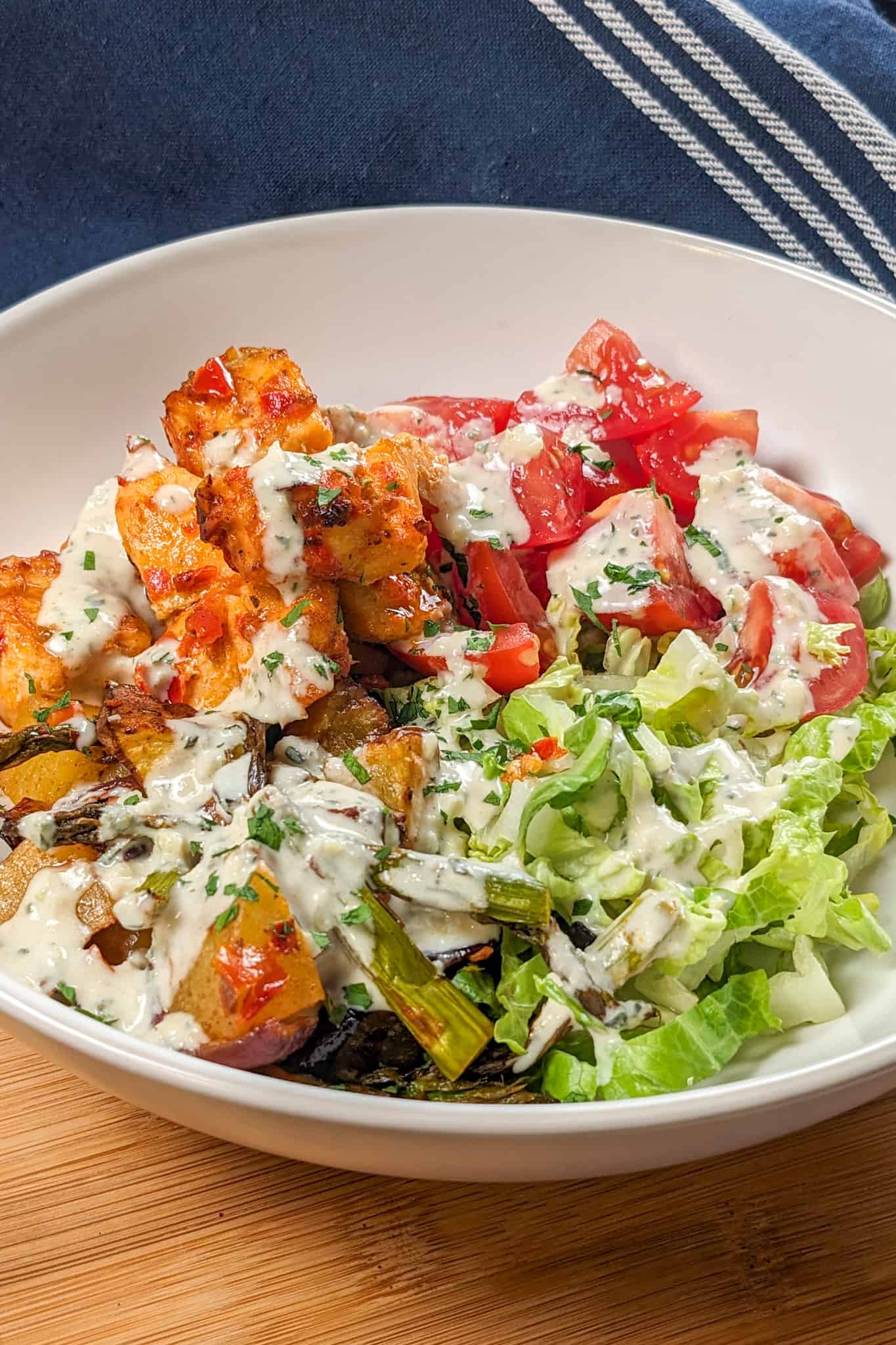 Side view of the Air Fryer Calabrian Chili Salmon and Vegetables Bowl in a wide rim bowl, a dish where the cubed baked salmon, quartered campari tomatoes, shredded romaine lettuce and roasted vegetables share equal pie part with the rosemary tahini dressing drizzled in a zig zag on top