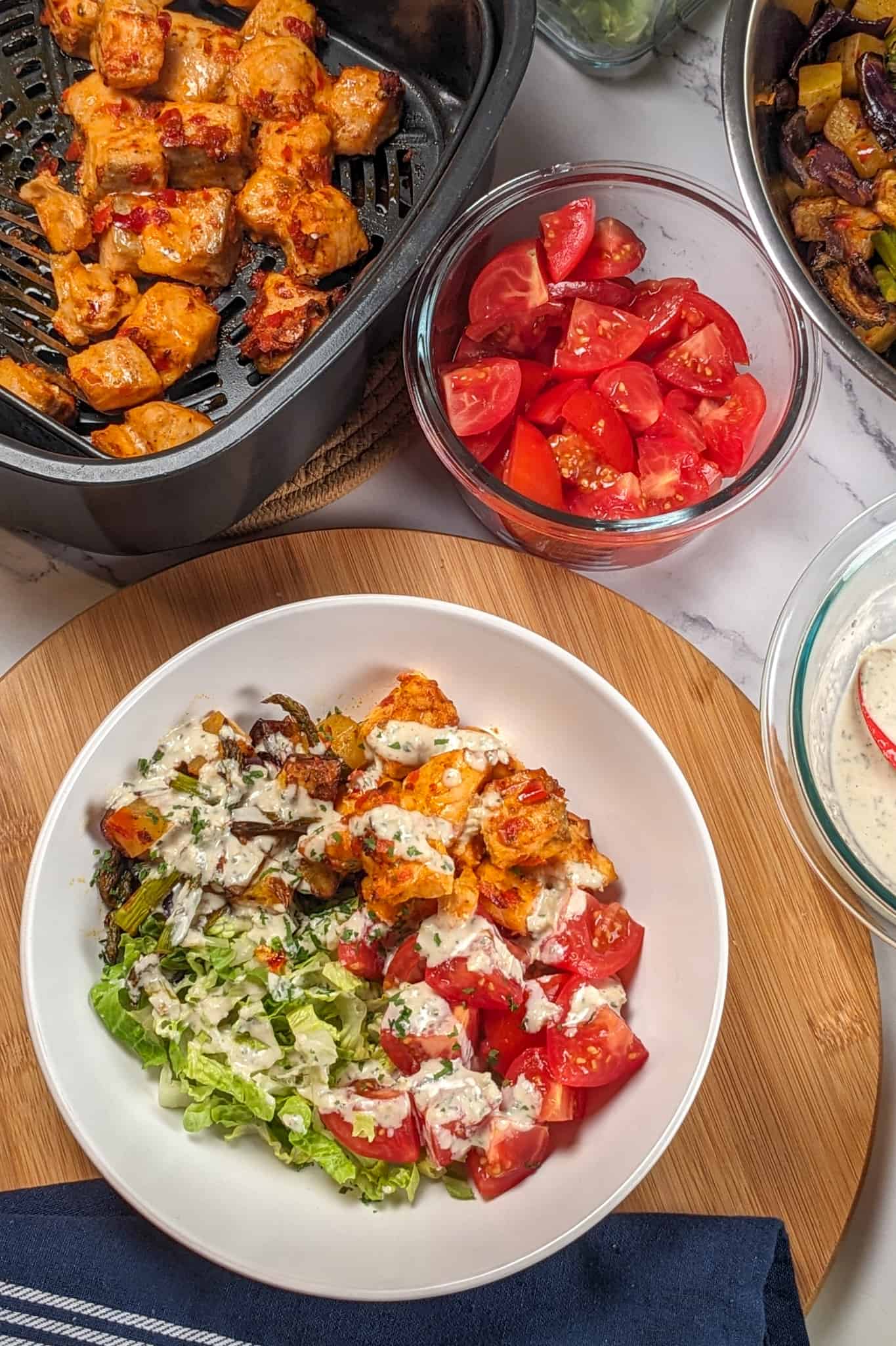 top view of the Air Fryer Calabrian Chili Salmon and Vegetables Bowl in a wide rim bowl, a dish where the cubed baked salmon, quartered campari tomatoes, shredded romaine lettuce and roasted vegetables share equal pie part with the rosemary tahini dressing drizzled in a zig zag on top on wooden lazy susan, surrounded by a kitchen towel, the air fryer canister with the baked cubed salmon, a round glass container of cut tomatoes, next to a stainless steel bowl of vegetables and another round glass container with the tahini dressing