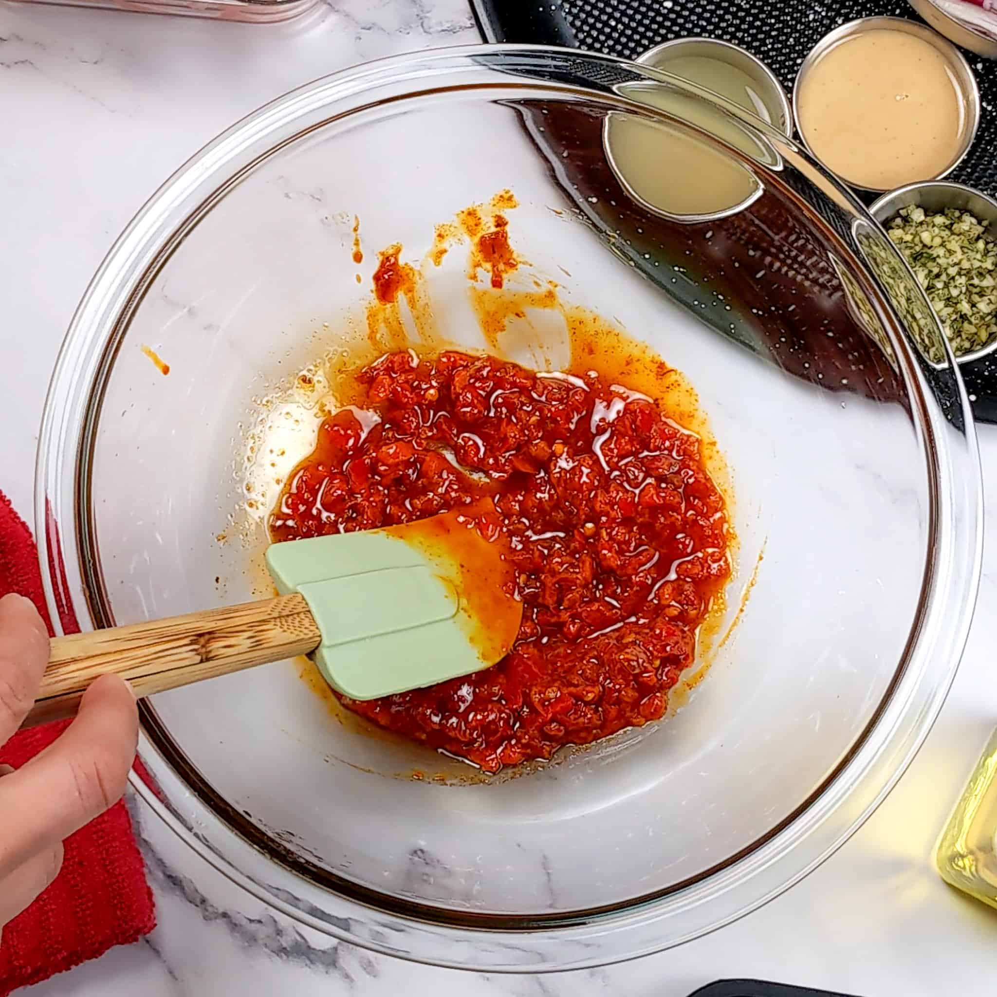 The calabrian chili wet rub in a pyrex glass mixing bowl being mixed with a kitchenaid silicone spatula with a wooden handle