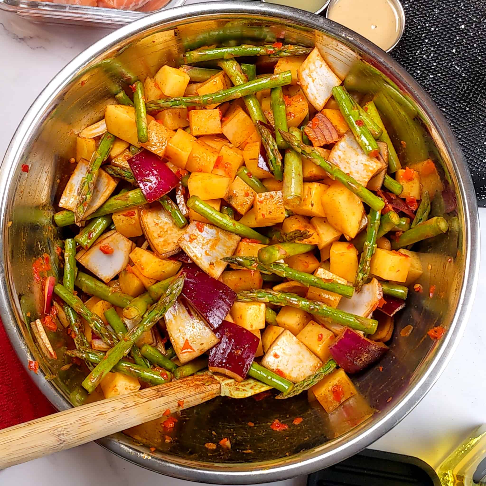 stainless steel bowl with the raw vegetables of large diced potatoes, cut asparagus and red onion in the calabrian chili wet rub with a silicone spatula tucked inside the mix.