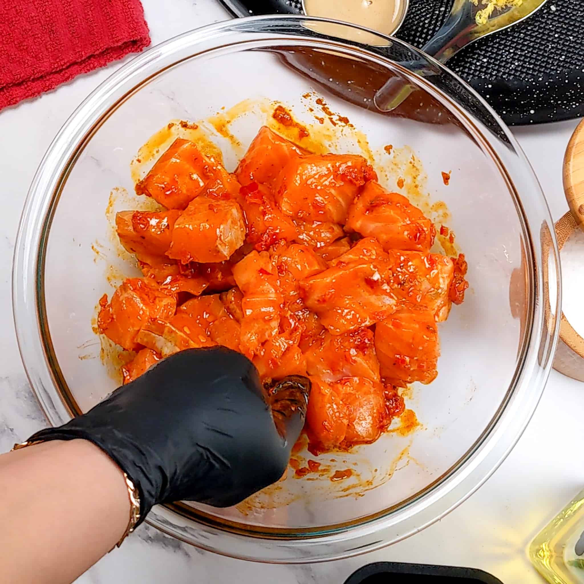 gloved hands mixing the calabrian chili paste with the large salmon chunks in a glass mixing bowl