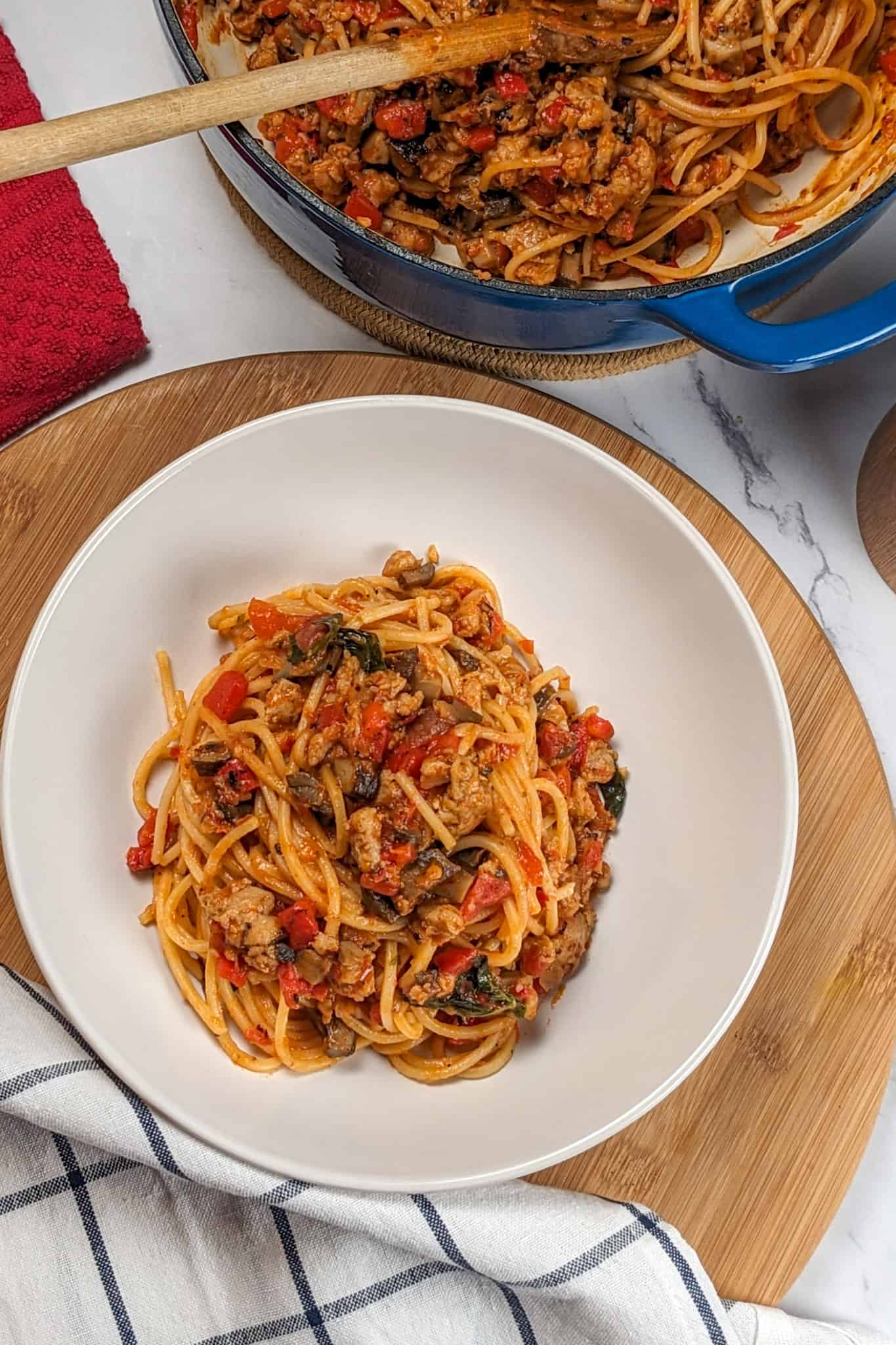 top view of the Easy Spicy Calabrian Italian Chicken Sausage Bolognese om a wide rim bowl on a wooden lazy susan, surrounded by a kitchen towels, and the dutch oven with the remaining pasta with a wooden spoon laying inside.