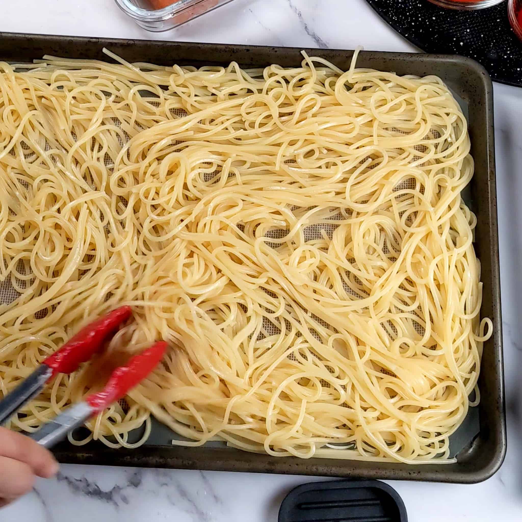 cooked spaghetti on a sheet pan lined with a silicone liner being spread out with a pair of kitchenaid silicone tipped tongs