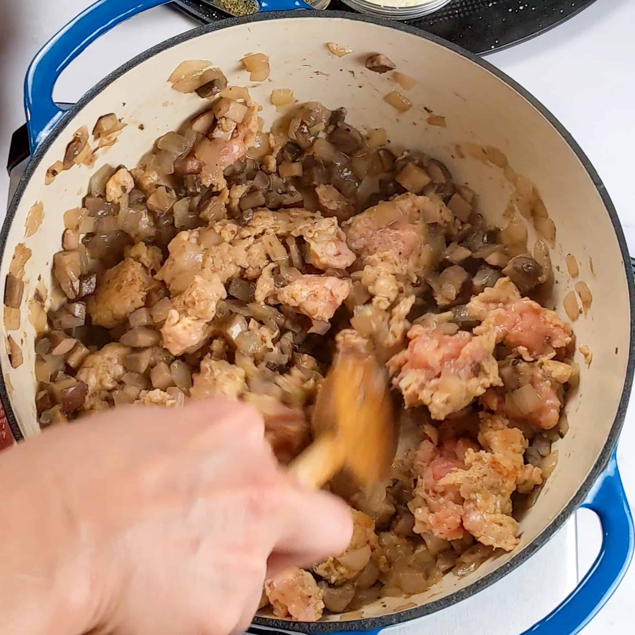 grounded chicken sausage being chopped wtih a wooden spoon with diced portobello mushrooms and onions sweating in a dutch oven