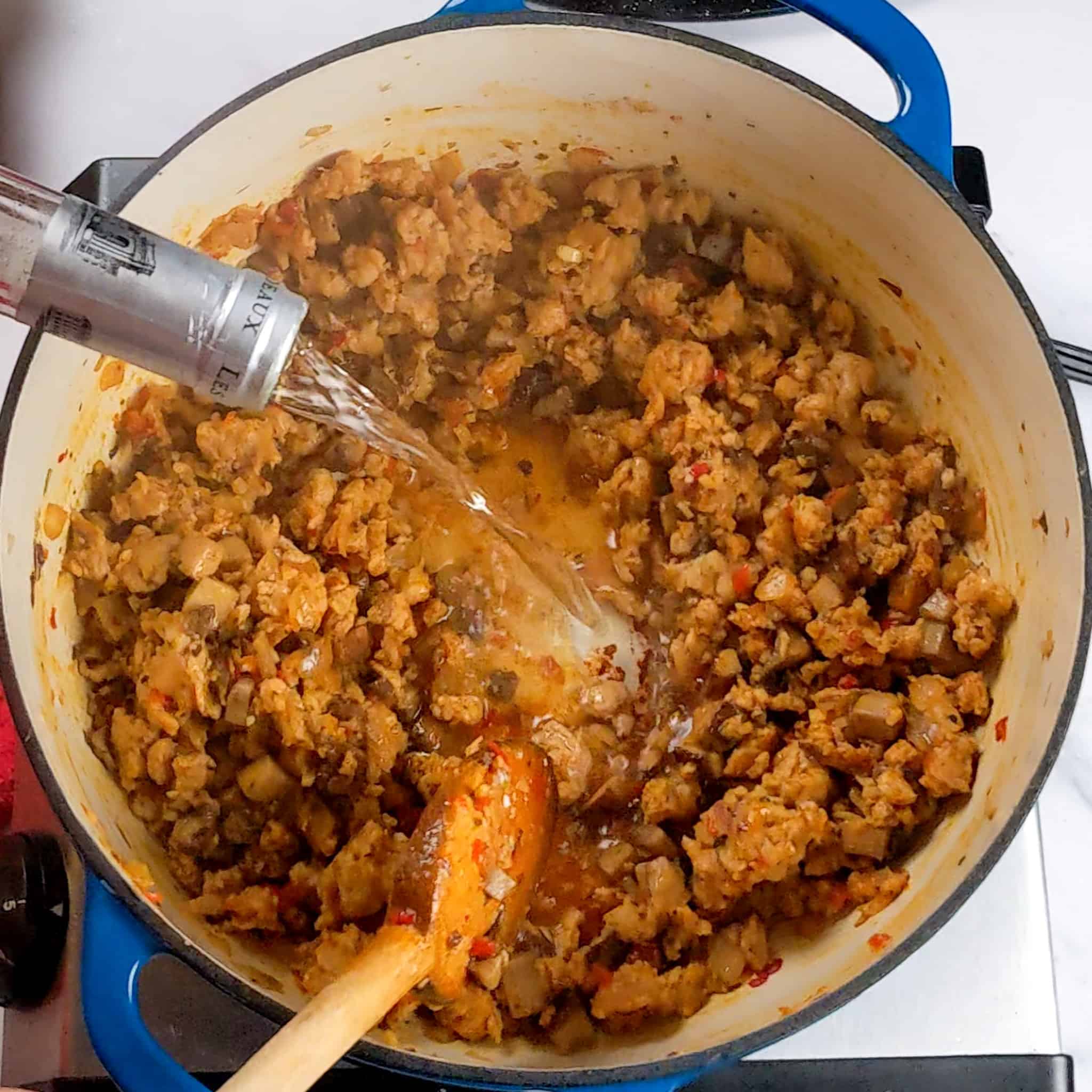 wine wine being poured into cooked crumbled sweet italian chicken sausage with diced portobello mushrooms and onions coated in crushed calabrian peppers in a dutch oven