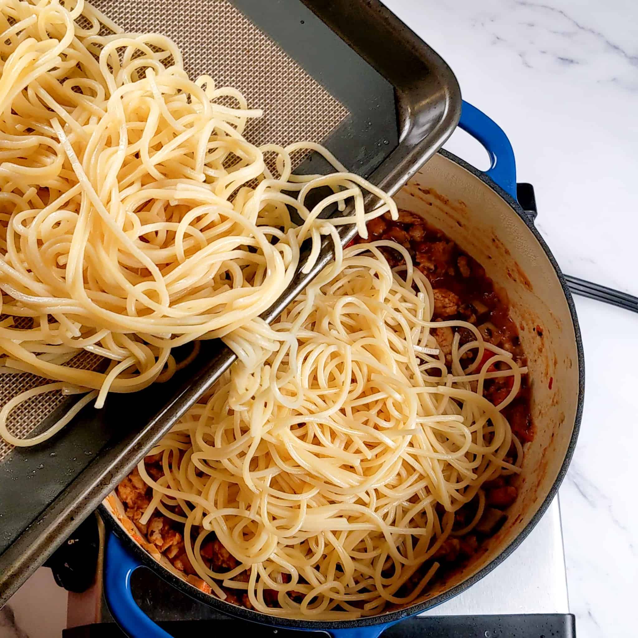 cooked cooled spaghetti on a baking sheet tray lined with a silicone liner being poured into the chicken sausage bolognese in a pot