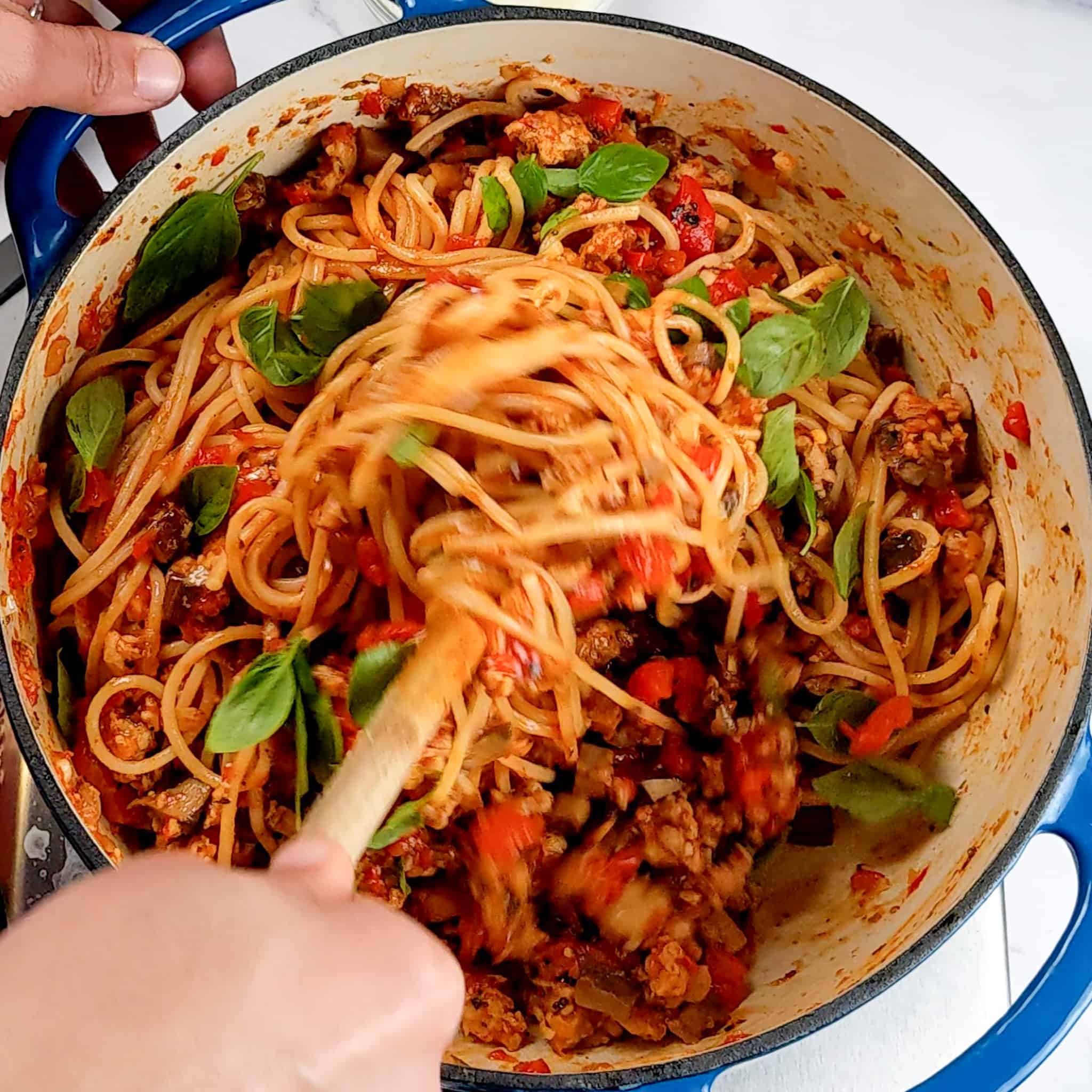 spaghetti being mixed into the chicken sausage bolognese in a pot with speckles of fresh basil leaves