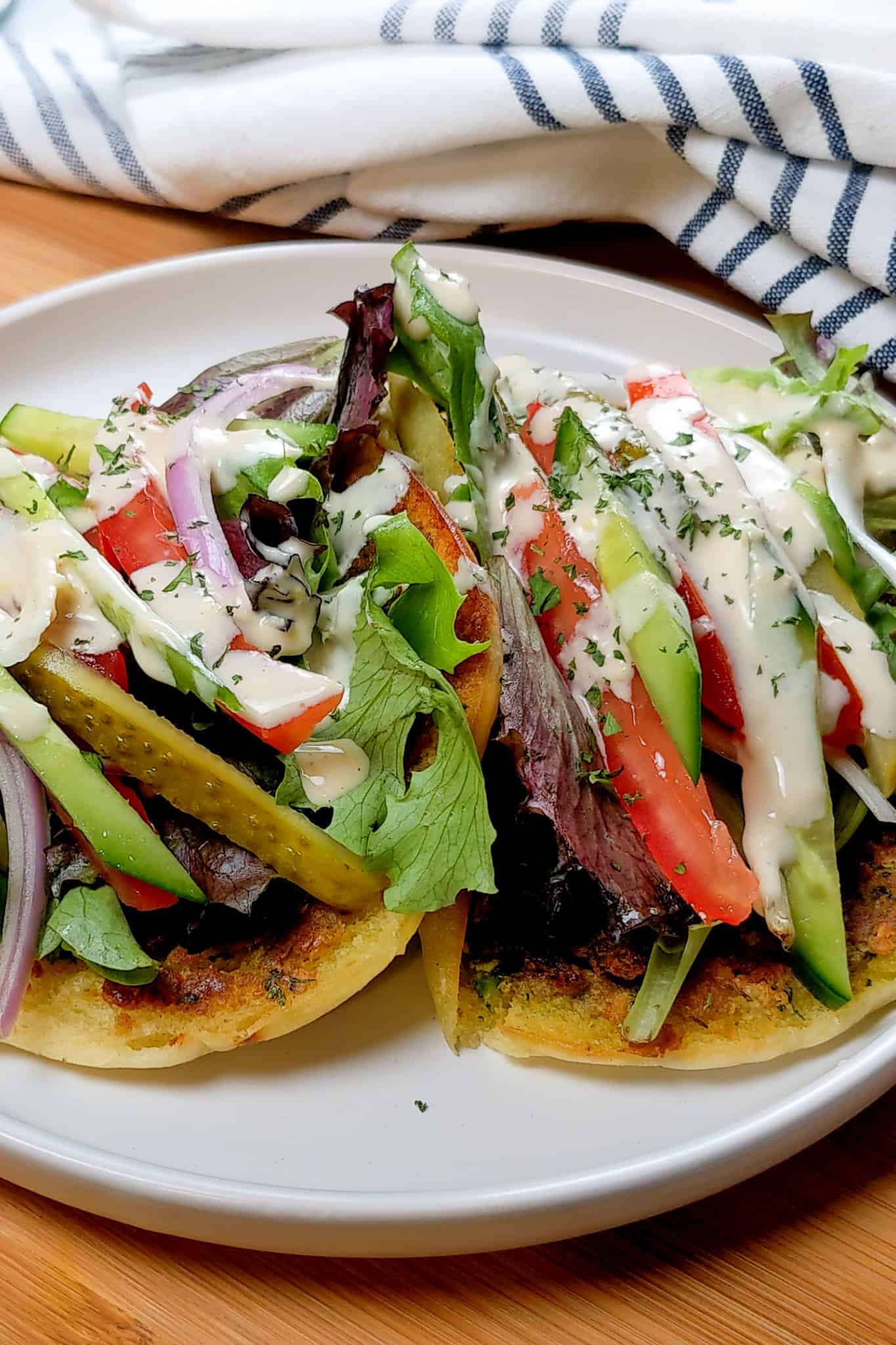 Side view of two Spicy Falafel Pita Sandwiches with Lemon Tahini Dressing on a round white plate on a wooden lazy susan with a kitchen towel in the background.